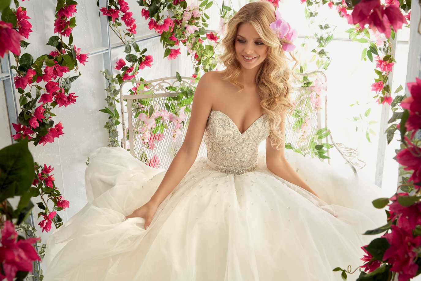 Wedding Dress 桌布 壁紙 — 一位光彩照人的新娘穿著華麗的白色禮服，在陽光普照的花園中，優雅地坐在粉紅色九重葛瀑布之間。