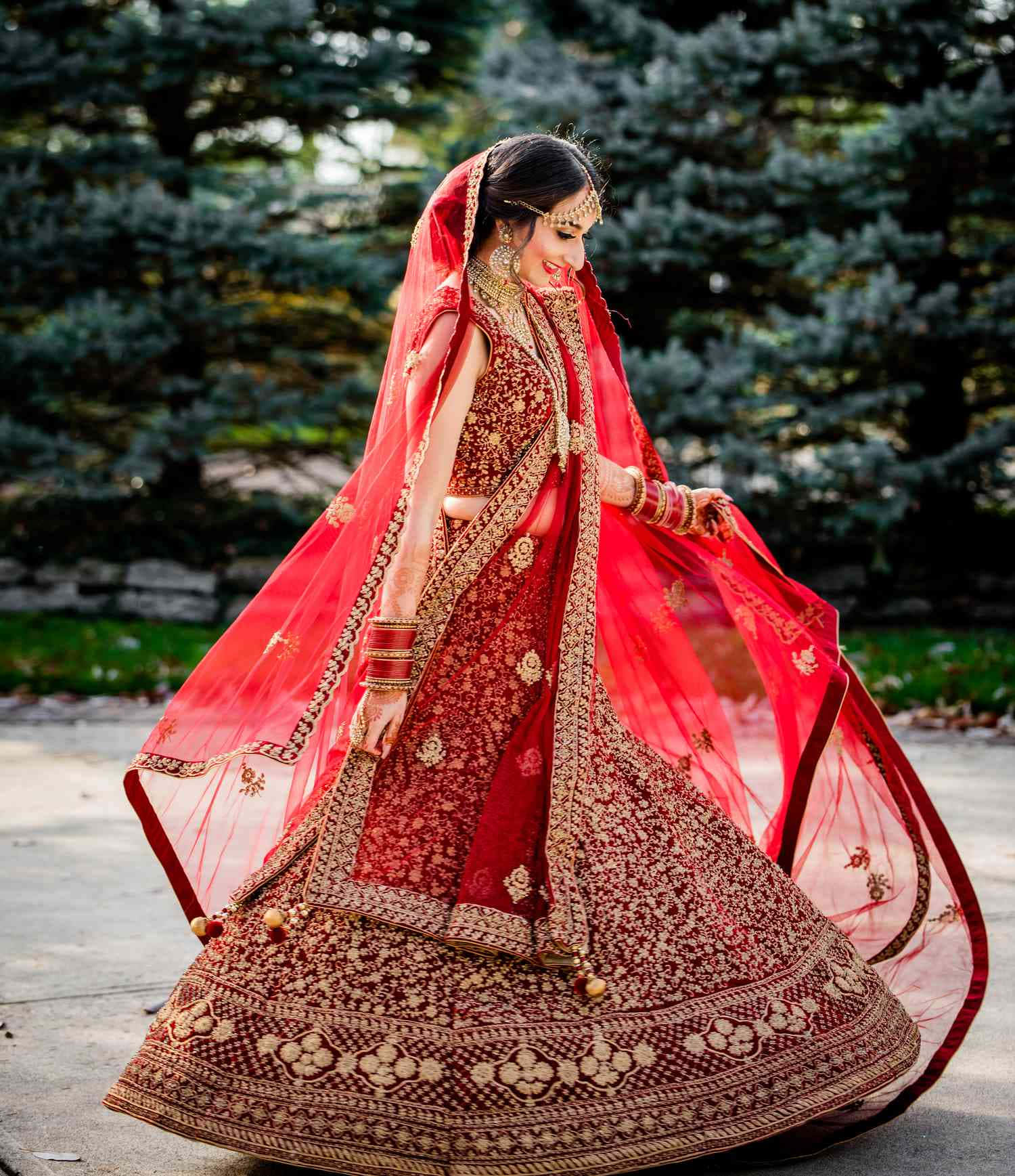 Bride 桌布 壁紙 — 一位光彩照人的新娘身穿華麗的紅金色刺繡 lehenga，在常青樹的背景下，面紗飄逸，優雅地在戶外旋轉。
