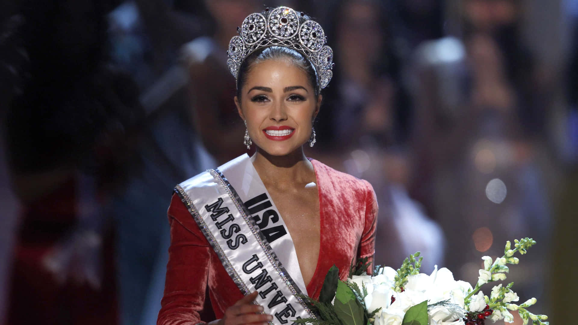 olivia culpo 桌布 壁紙 — 一位光彩照人的美國環球小姐冠軍，身穿紅色天鵝絨長裙，頭戴閃亮皇冠，在舞台上手持花束微笑。