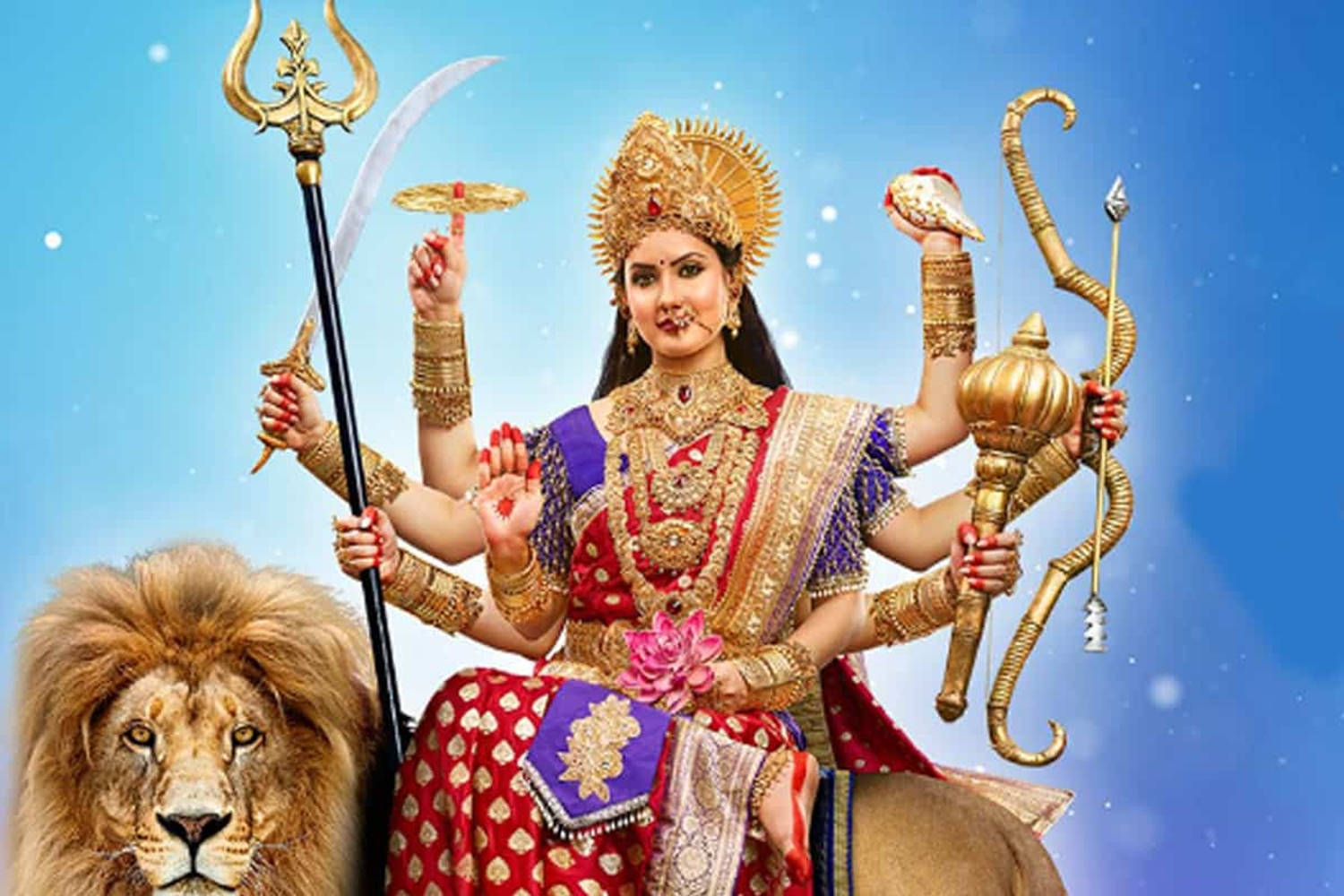 Vaishno Devi 桌布 壁紙 — 一位光芒四射的多臂女神杜爾迦，身著華麗的紅色和金色服飾，騎著獅子，背景是閃爍的藍天。