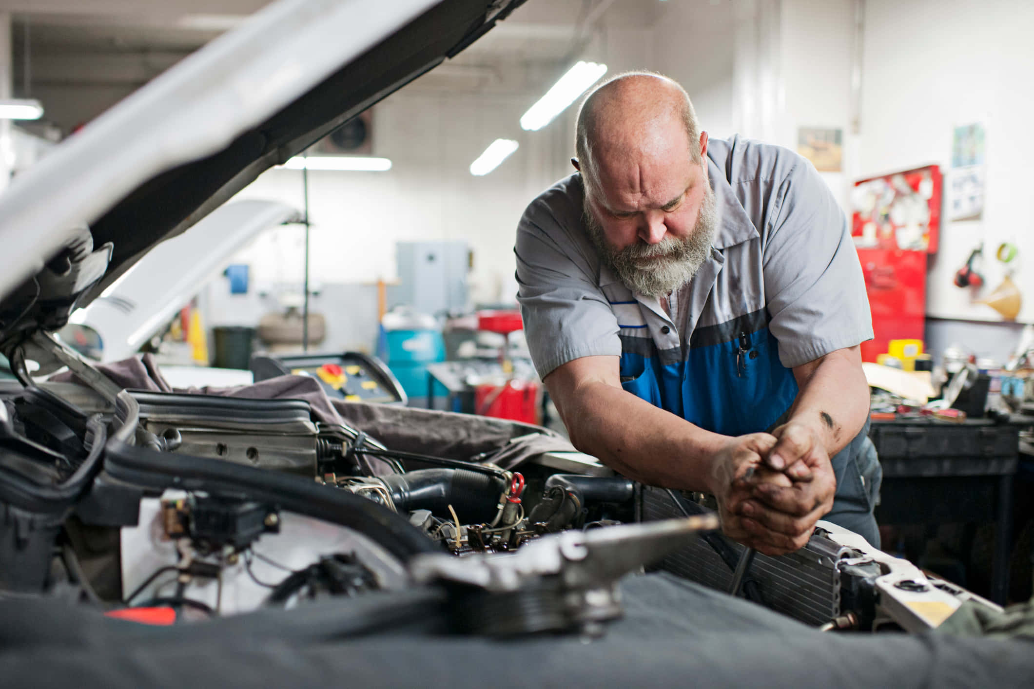 Car Repair 桌布 壁紙 — 一位光頭、留著灰色鬍鬚的機械師在光線充足、工具齊全的車庫裡專注地修理汽車引擎蓋下的零件。
