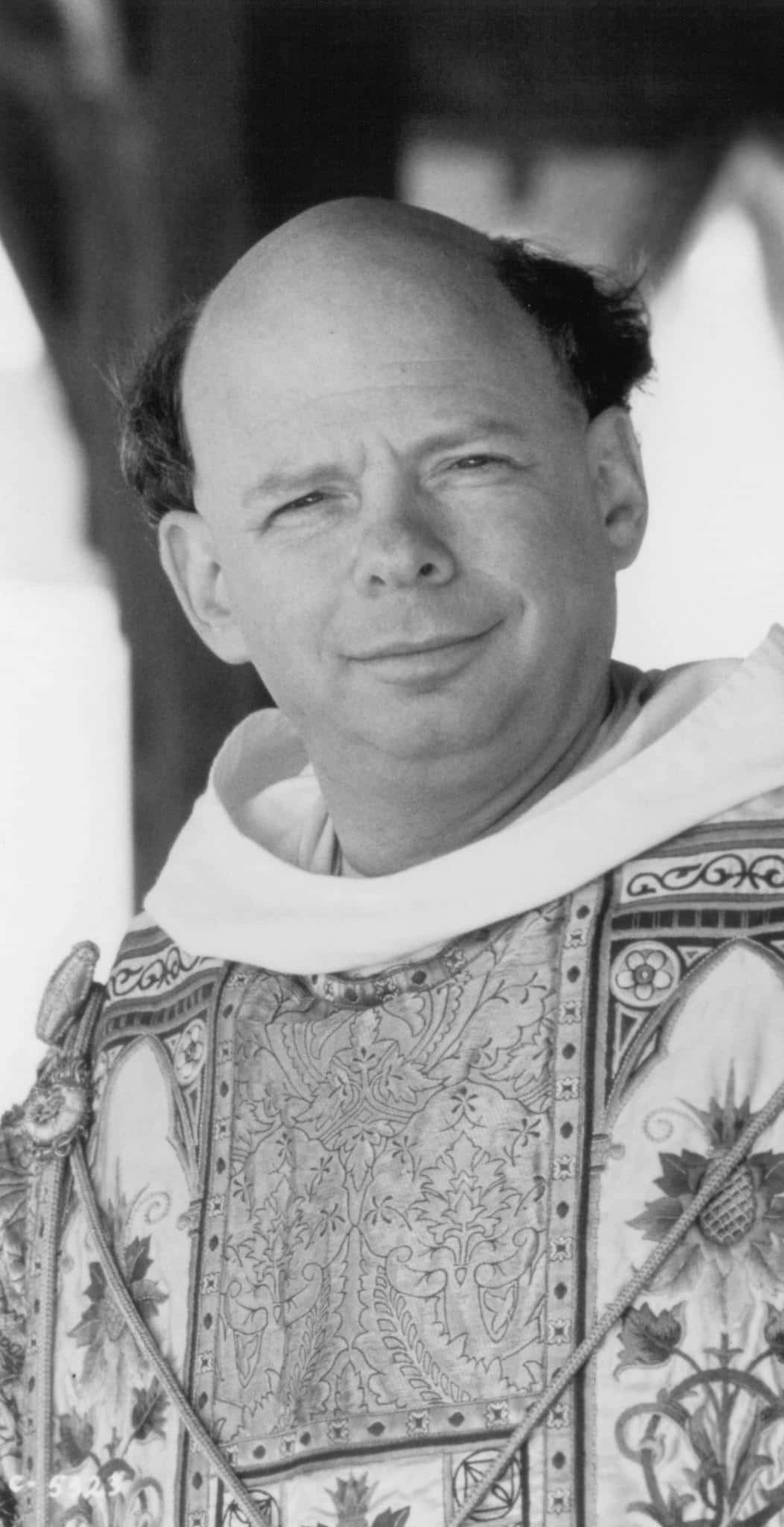 Wallace Shawn 桌布 壁紙 — 一位光頭男士的黑色和白色肖像，面帶微笑，穿著帶有花卉和幾何刺繡的華麗儀式長袍。