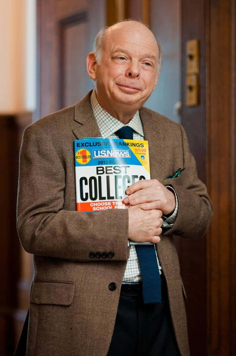 Wallace Shawn 桌布 壁紙 — 一位光頭男士身穿棕色粗花呢外套，在木鑲板走廊中自信微笑，手持US News Best Colleges雜誌。