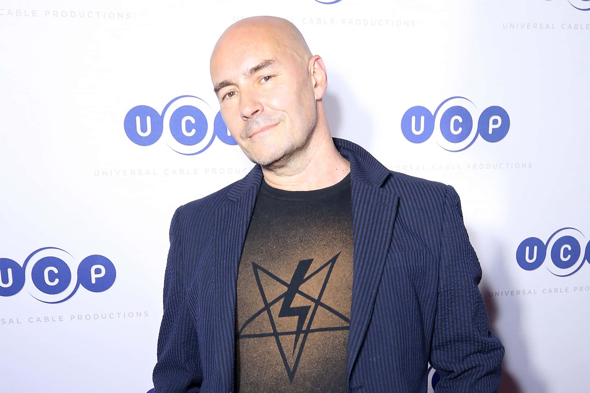 Grant Morrison 桌布 壁紙 — 一位光頭男士身穿海軍藍細條紋西裝外套和圖案T恤，在品牌活動背景前對著鏡頭微笑。