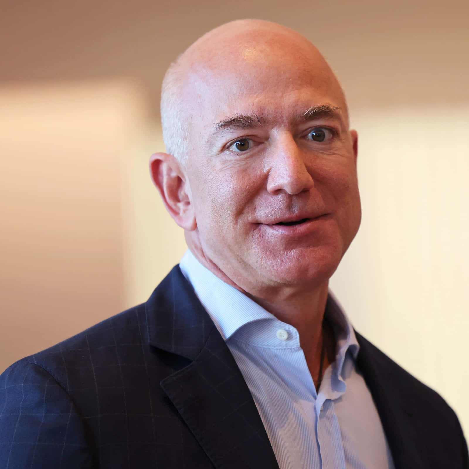 Jeff Bezos 桌布 壁紙 — 一位光頭男士身穿深色格紋西裝和淺藍色襯衫，在柔和的米色背景下溫暖微笑的特寫肖像。