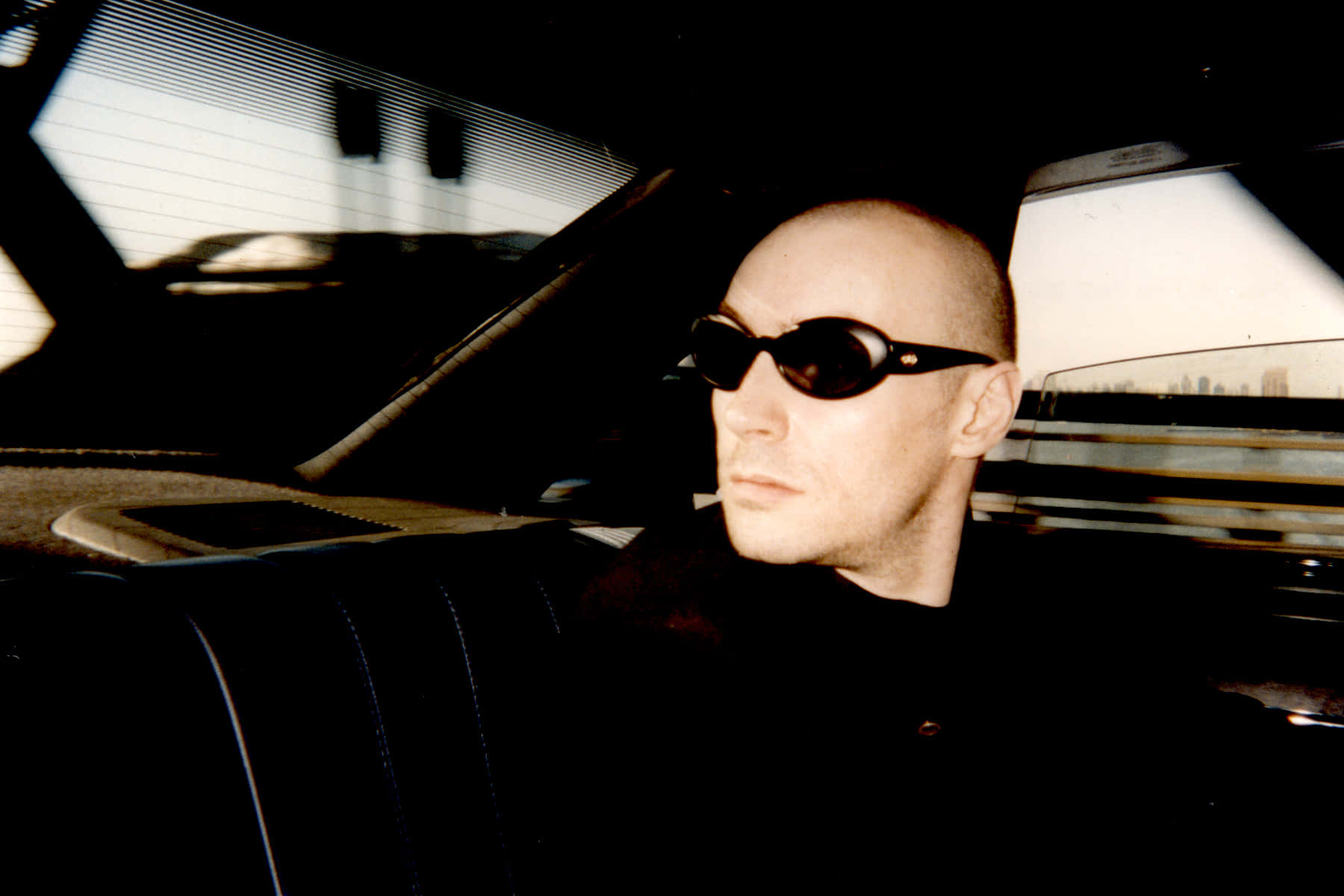 Grant Morrison 桌布 壁紙 — 一位光頭男子戴著深色太陽眼鏡，靜靜地坐在一輛復古車內，車內有陽光照射的百葉窗和城市景觀的倒影。
