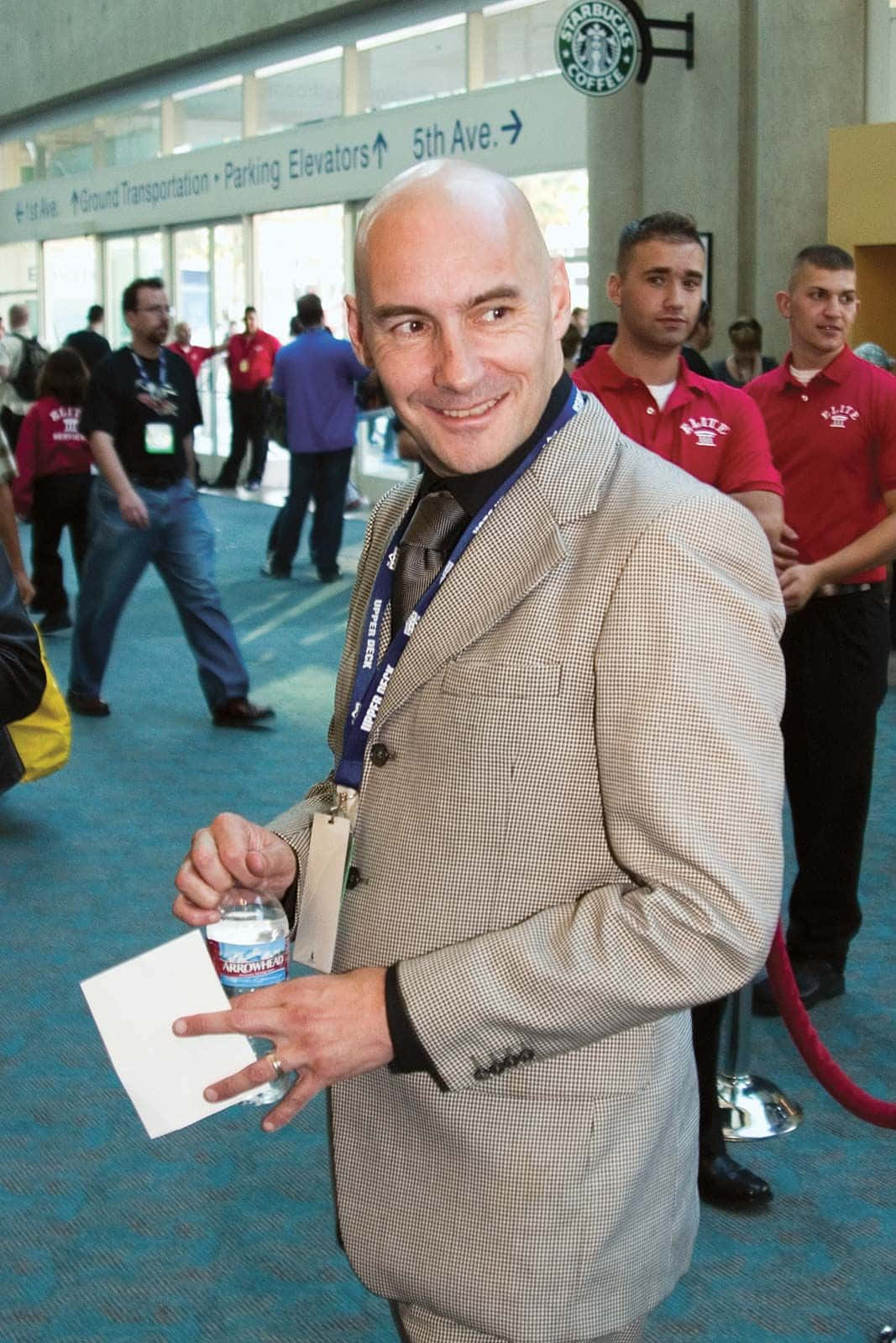 Grant Morrison 桌布 壁紙 — 一位光頭男子穿著格紋西裝，在繁忙的會議中心微笑著拿著水瓶。