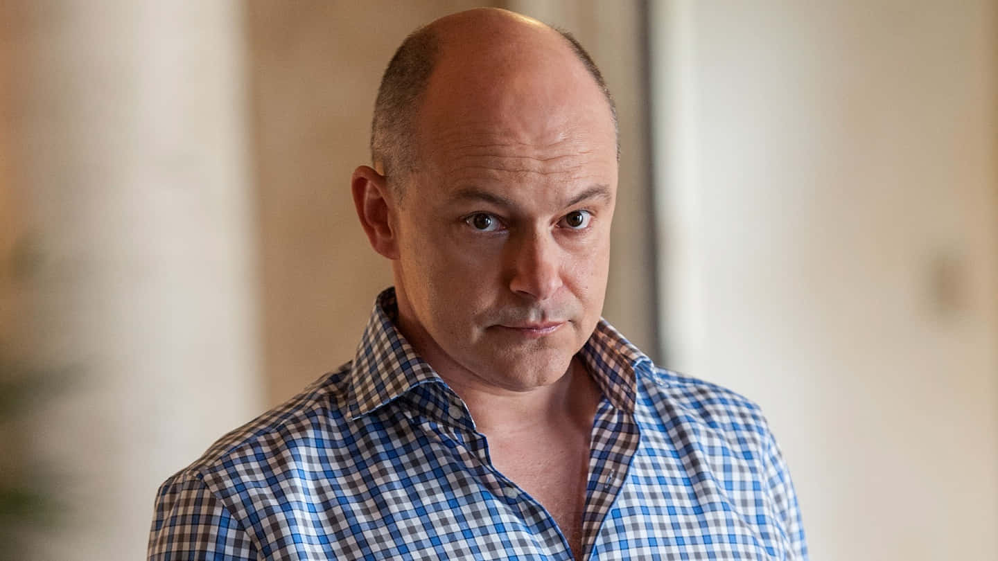 Rob Corddry 桌布 壁紙 — 一位光頭男子身穿藍色格仔襯衫，在室內以嚴肅的表情專注地凝視。