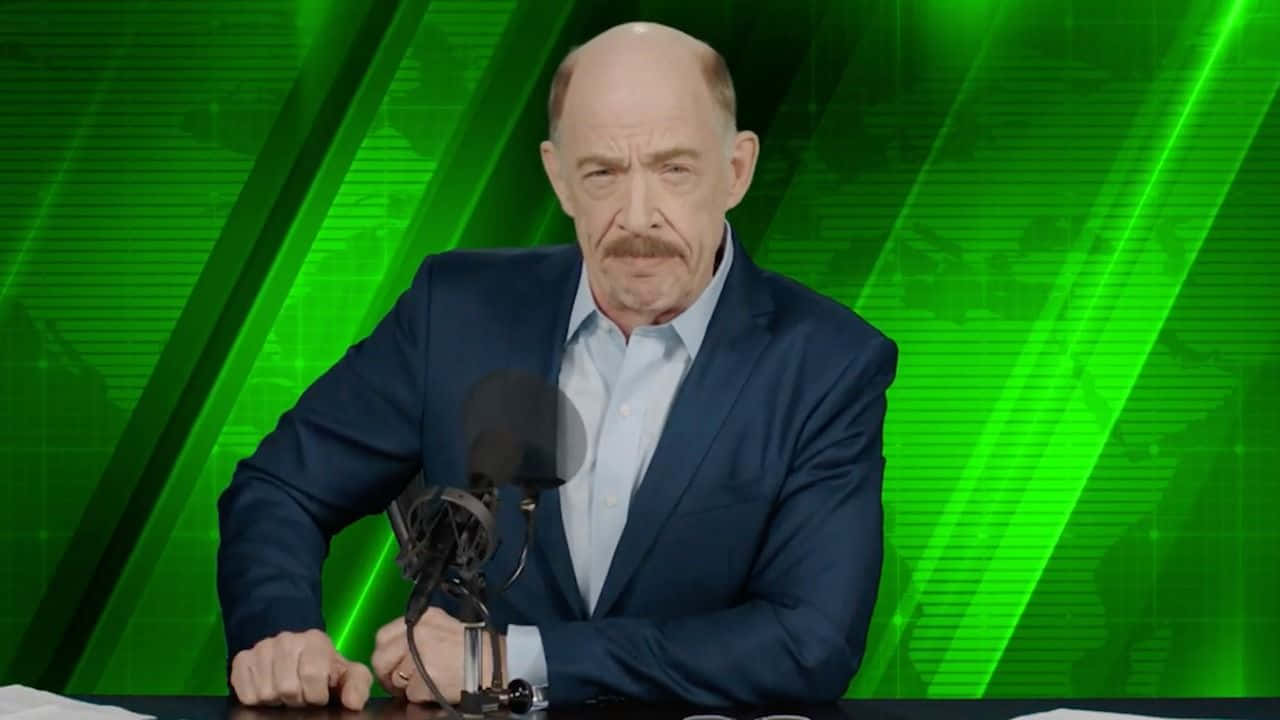 J Jonah Jameson 桌布 壁紙 — 一位光頭留鬍鬚的男人穿著藍色西裝，坐在有咪高峰的桌前，背景是充滿活力的綠色數碼背景。