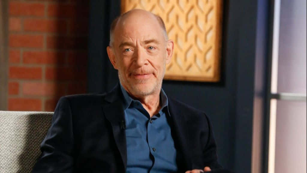 J Jonah Jameson 桌布 壁紙 — 一位光頭鬍鬚男，身穿深色西裝外套和藍色襯衫，坐在椅子上，背景是磚牆和圖案藝術品。