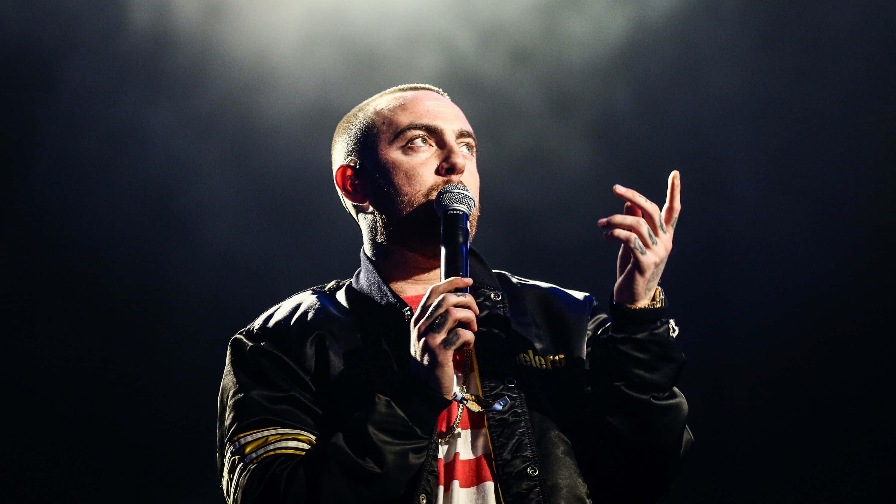 Mac Miller 桌布 壁紙 — 一位光頭鬍鬚男在黑暗背景下，於戲劇性聚光燈下拿著麥克風在舞台上表演。