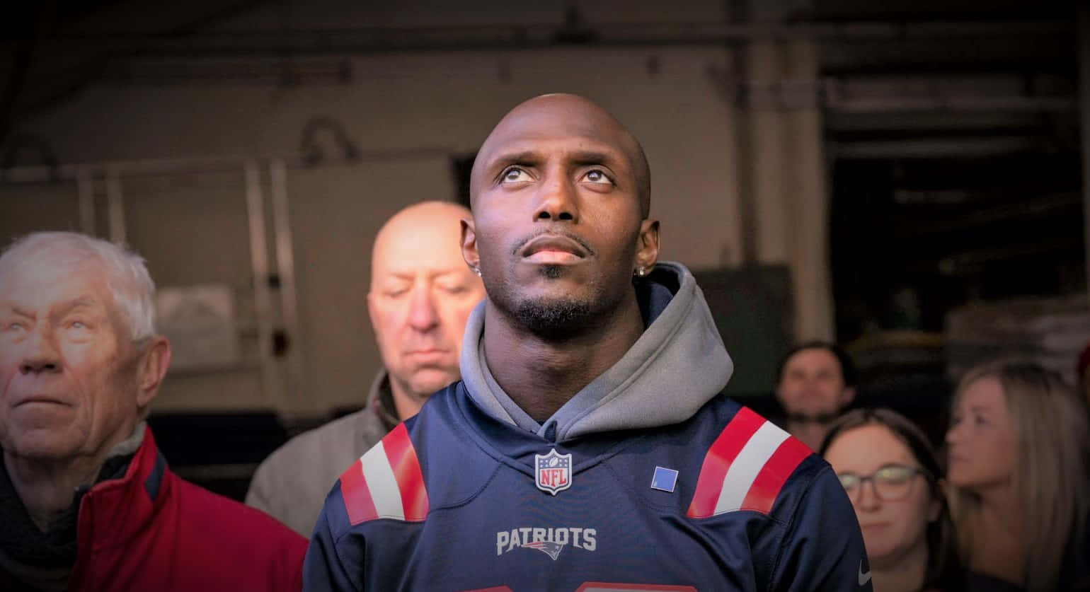 Devin Mccourty 桌布 壁紙 — 一位光頭黑人男子身穿Patriots NFL連帽衫，在昏暗的室內環境中與其他人坐在一起，表情嚴肅地向上凝視。