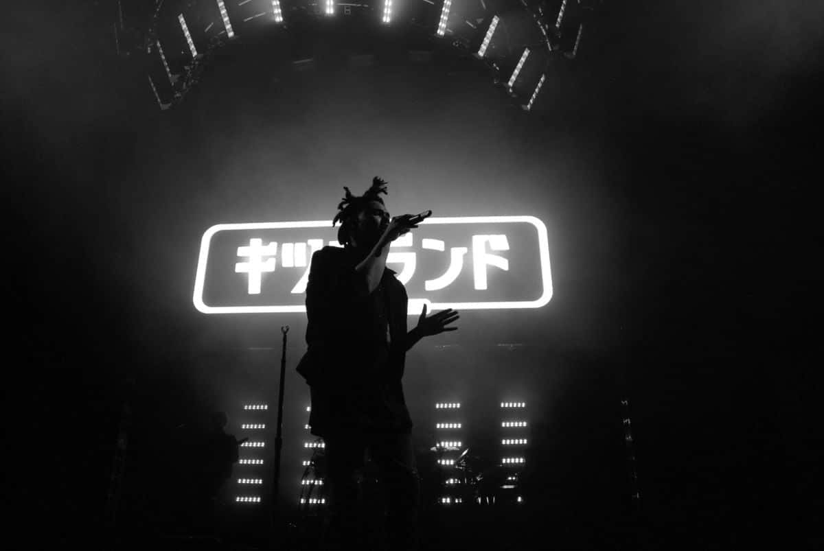 The Weeknd 桌布 壁紙 — 一位剪影表演者在黑暗的舞台上，在發光的日文字和聚光燈下說唱。