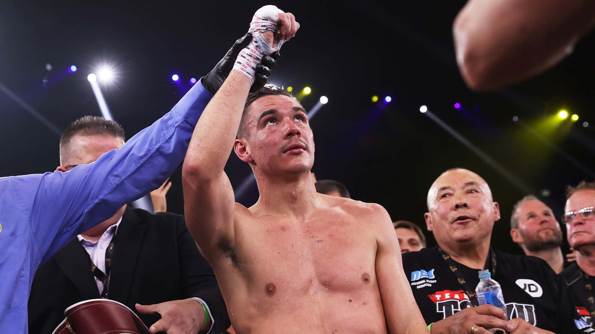 Tim Tszyu 桌布 壁紙 — 一位勝利的拳擊手在燈光明亮的擂台上被裁判舉起手臂，周圍是觀眾。