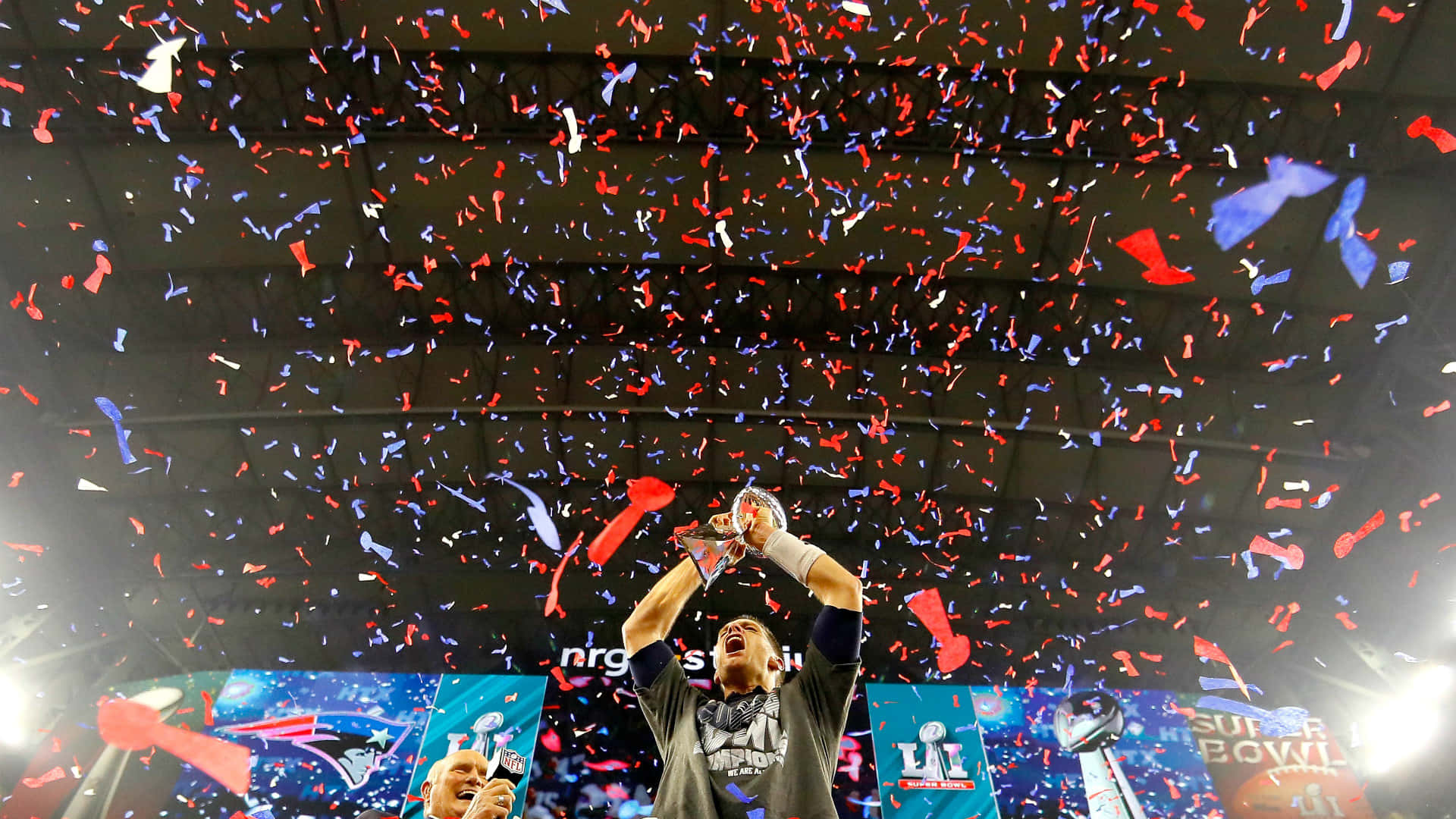 Super Bowl 桌布 壁紙 — 一位勝利的足球員在體育館內高舉超級碗獎盃，紅色、藍色和白色的紙屑在他周圍爆炸。