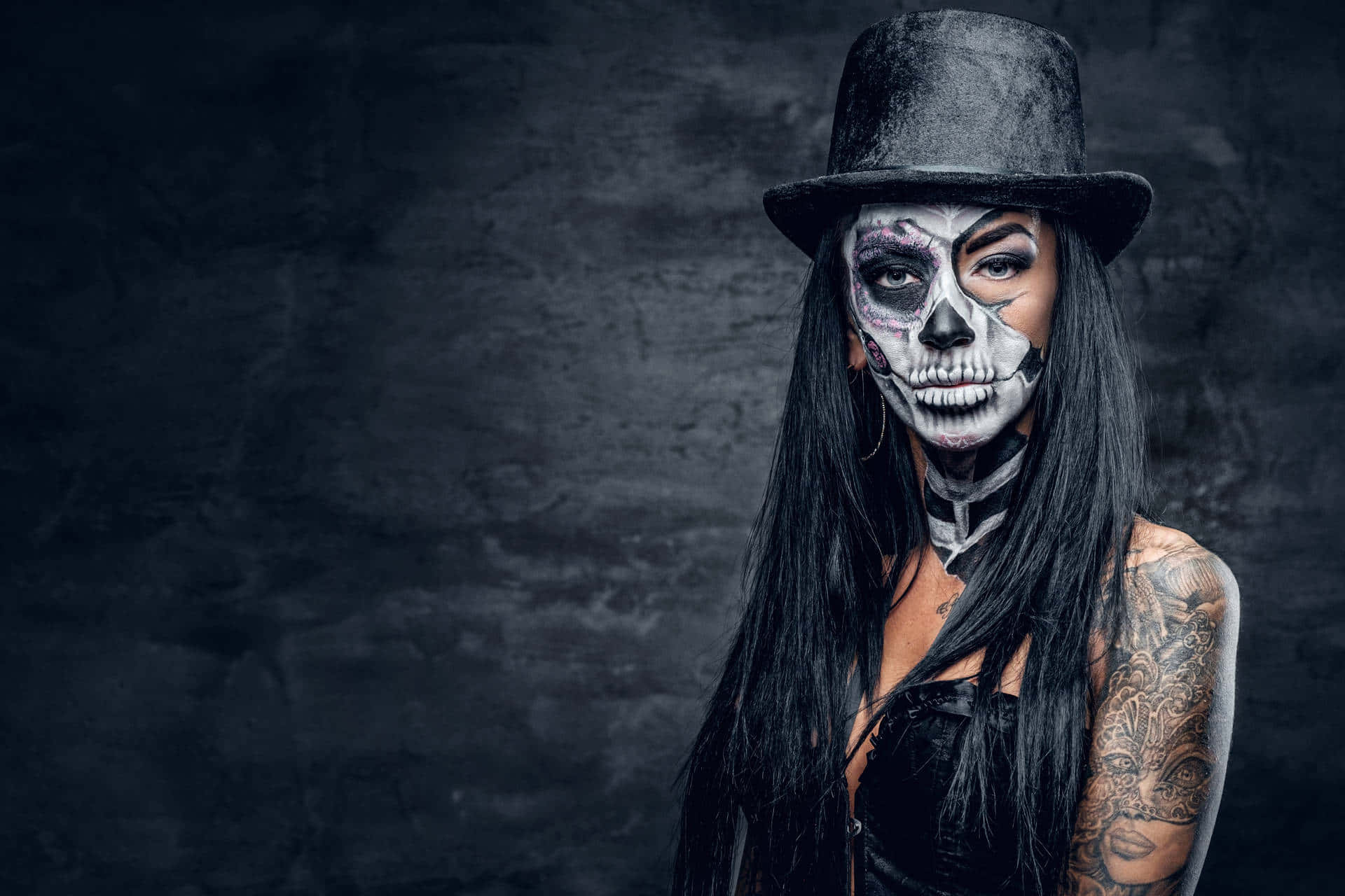 Halloween Tattoos 桌布 壁紙 — 一位化著半骷髏妝容及紋身的女性，戴著黑色高帽，在深色紋理牆前擺姿勢。