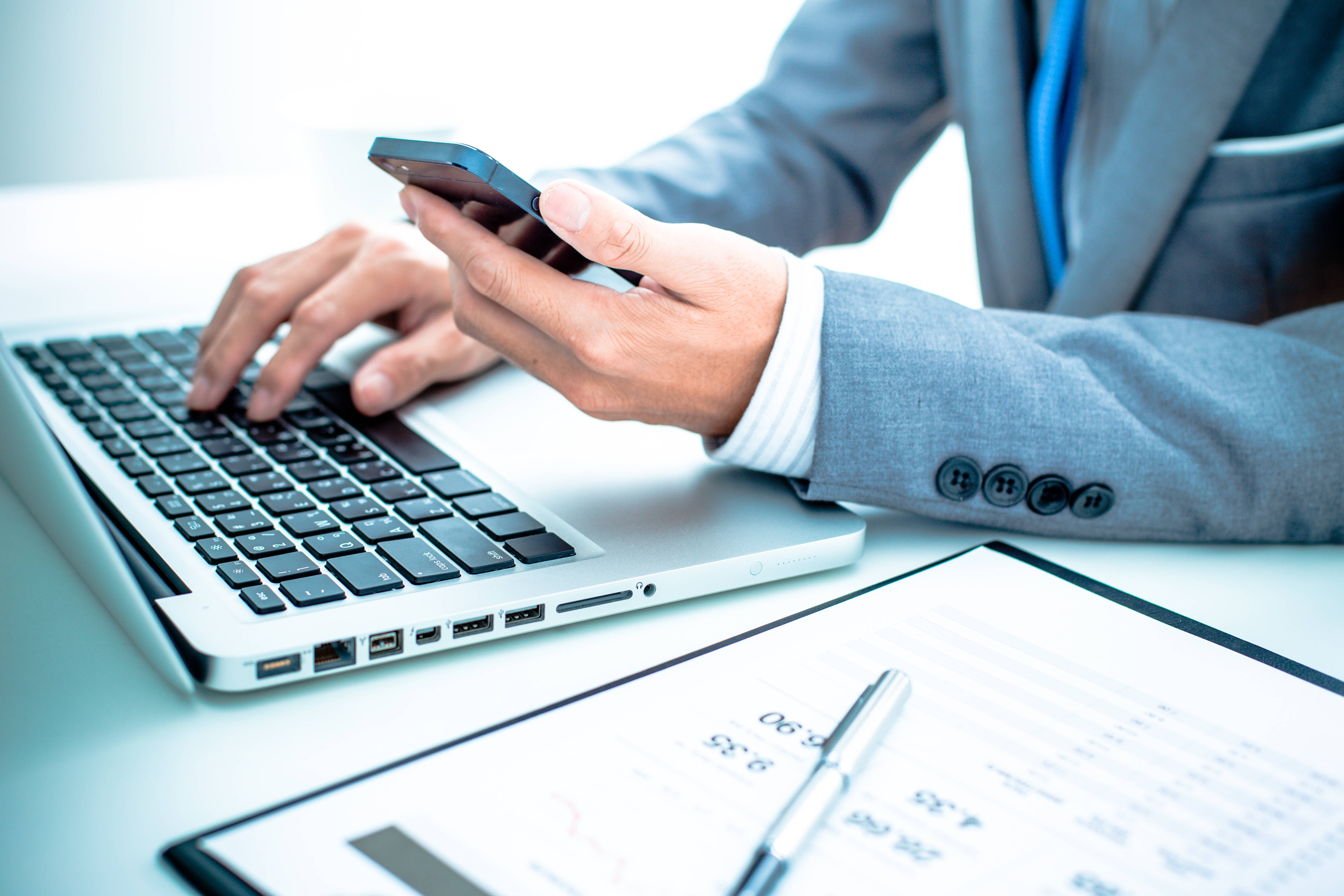 Chartered Accountant 桌布 壁紙 — 一位商人在辦公桌上使用手提電腦和智能手機同時處理多項任務，並審閱文件。