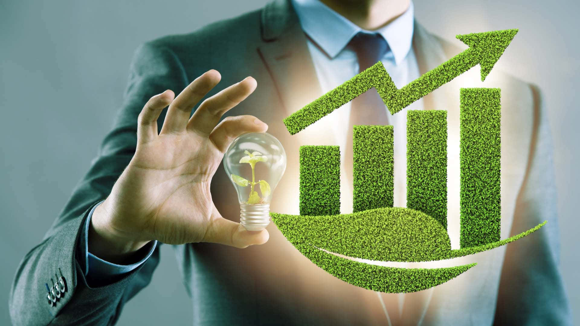 Green Economy 桌布 壁紙 — 一位商人手持一個發光的燈泡，裏面有嫩芽，旁邊漂浮著一個草質的上升條形圖和一片葉子。