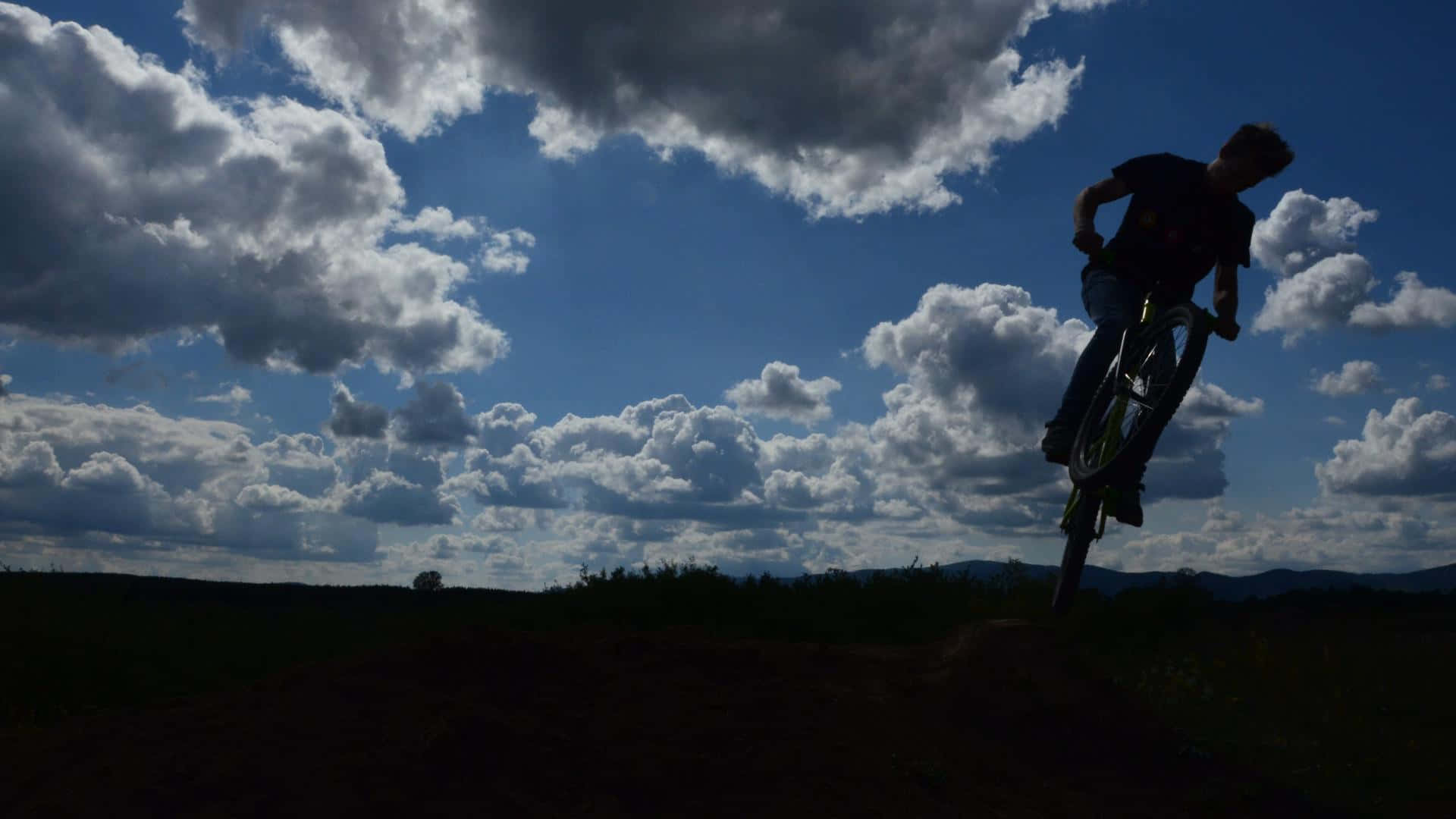 Mtb 桌布 壁紙 — 一位單車手的剪影在戲劇性的多雲天空下，於開闊田野中跳過一個土丘。