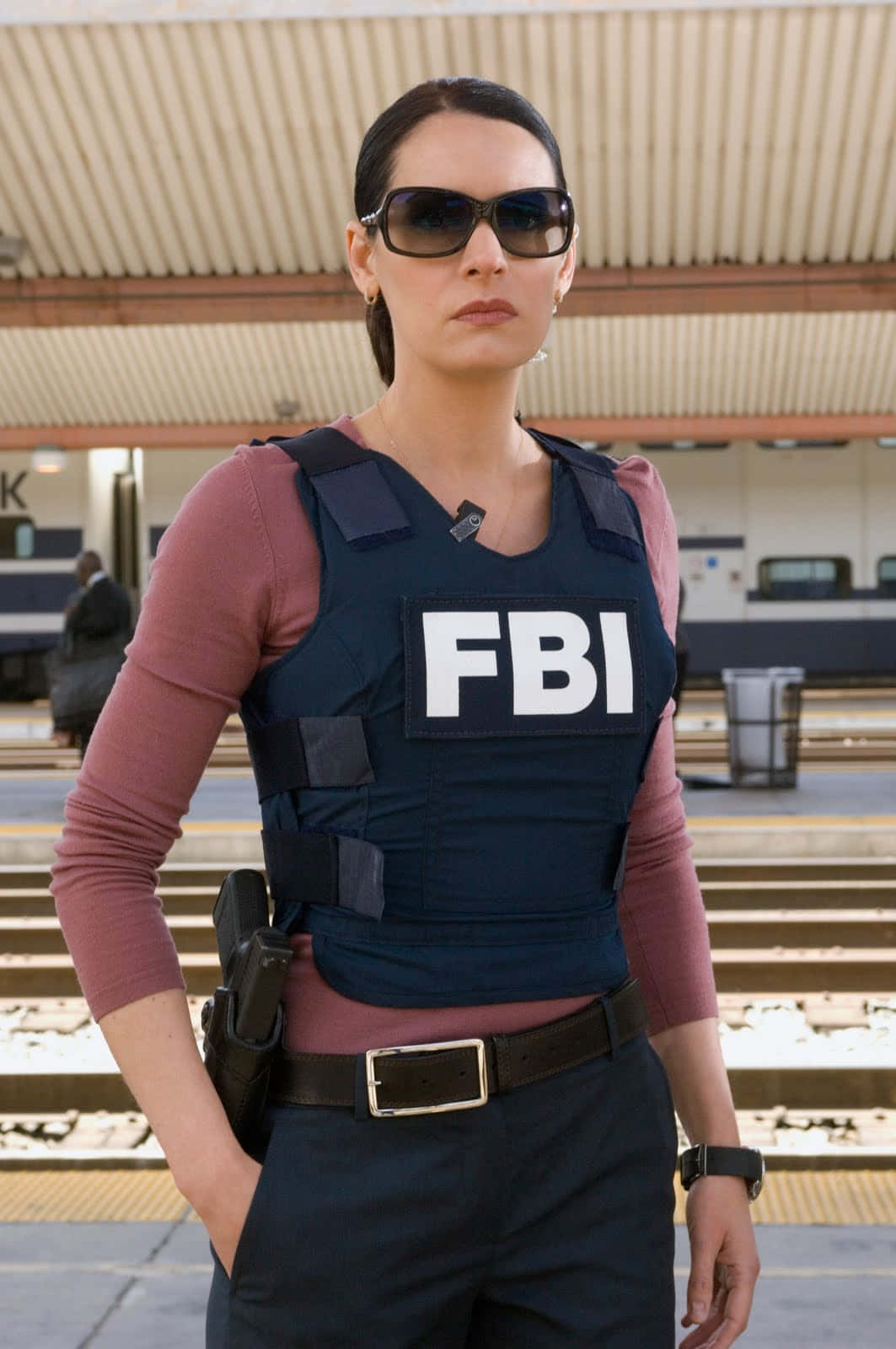paget brewster 桌布 壁紙 — 一位嚴肅的女FBI探員戴著太陽眼鏡和戰術背心，自信地站在火車站月台上。