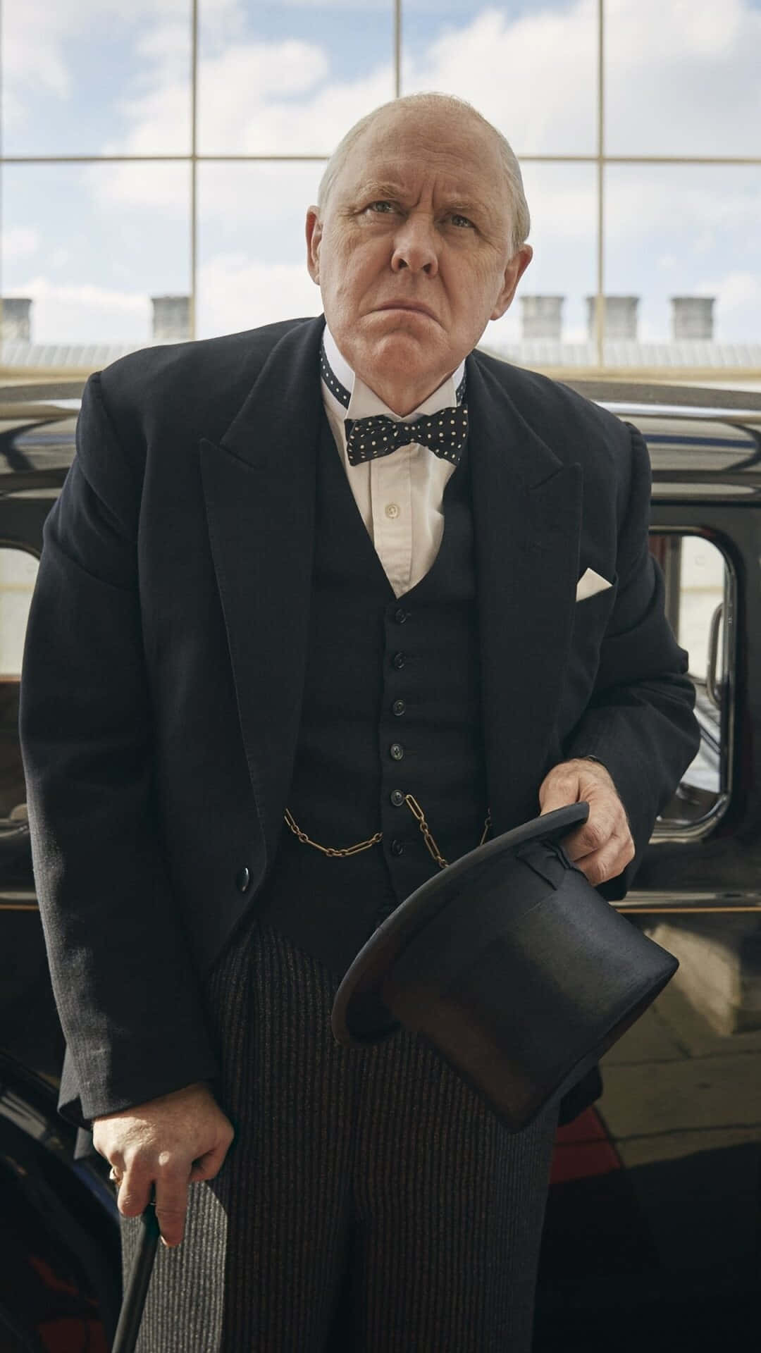 John Lithgow 桌布 壁紙 — 一位嚴肅的老年男子穿著正式黑色西裝和領結，站在一輛老爺車旁，手持高頂禮帽和手杖。