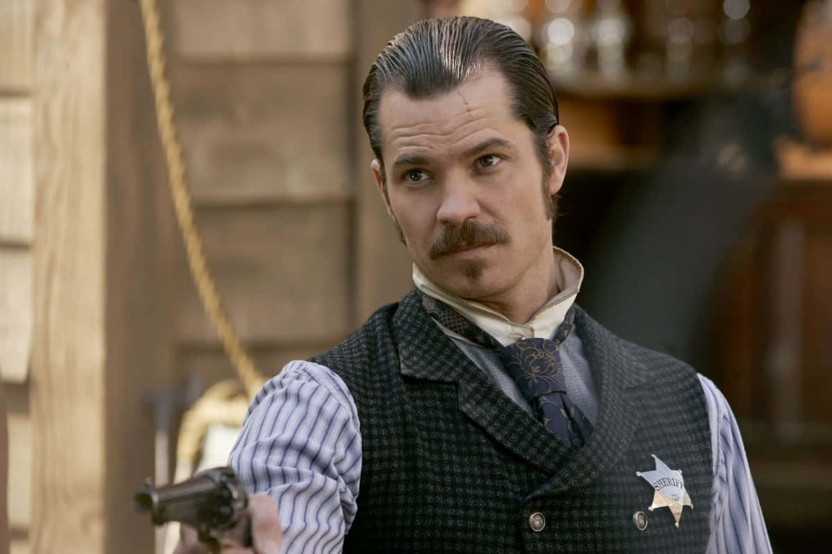 timothy olyphant 桌布 壁紙 — 一位嚴肅的警長留著鬍子，在塵土飛揚的西部沙龍中握著一把左輪手槍。