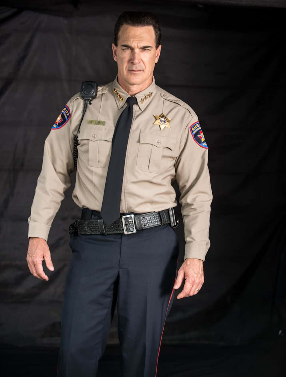 Patrick Warburton 桌布 壁紙 — 一位嚴肅的警長穿著米色制服，站在黑色背景前，表情嚴肅，徽章清晰可見。