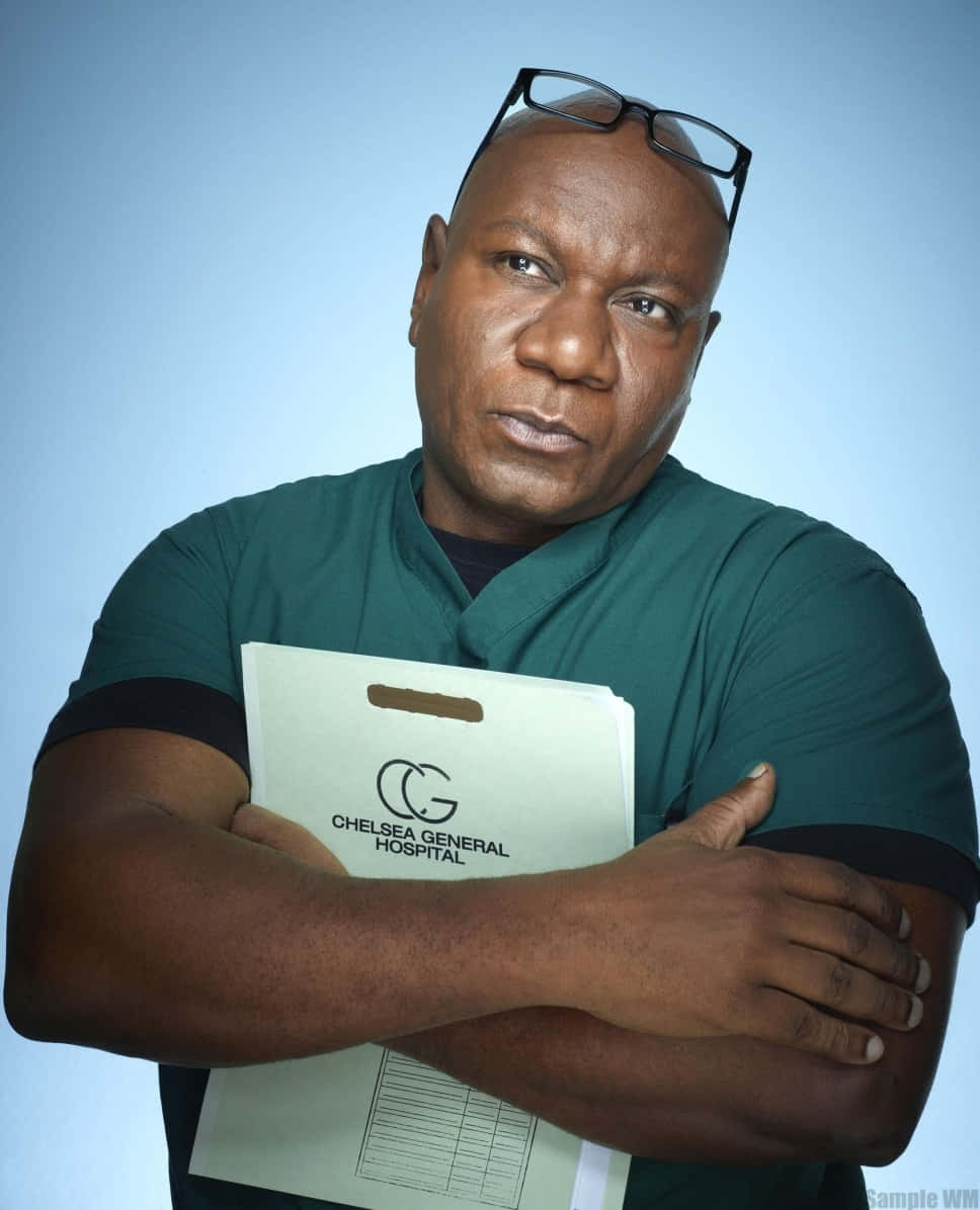 Ving Rhames 桌布 壁紙 — 一位嚴肅的黑人男醫生穿著綠色手術服，在藍色攝影棚背景前交叉雙臂，拿著Chelsea General Hospital的文件夾。