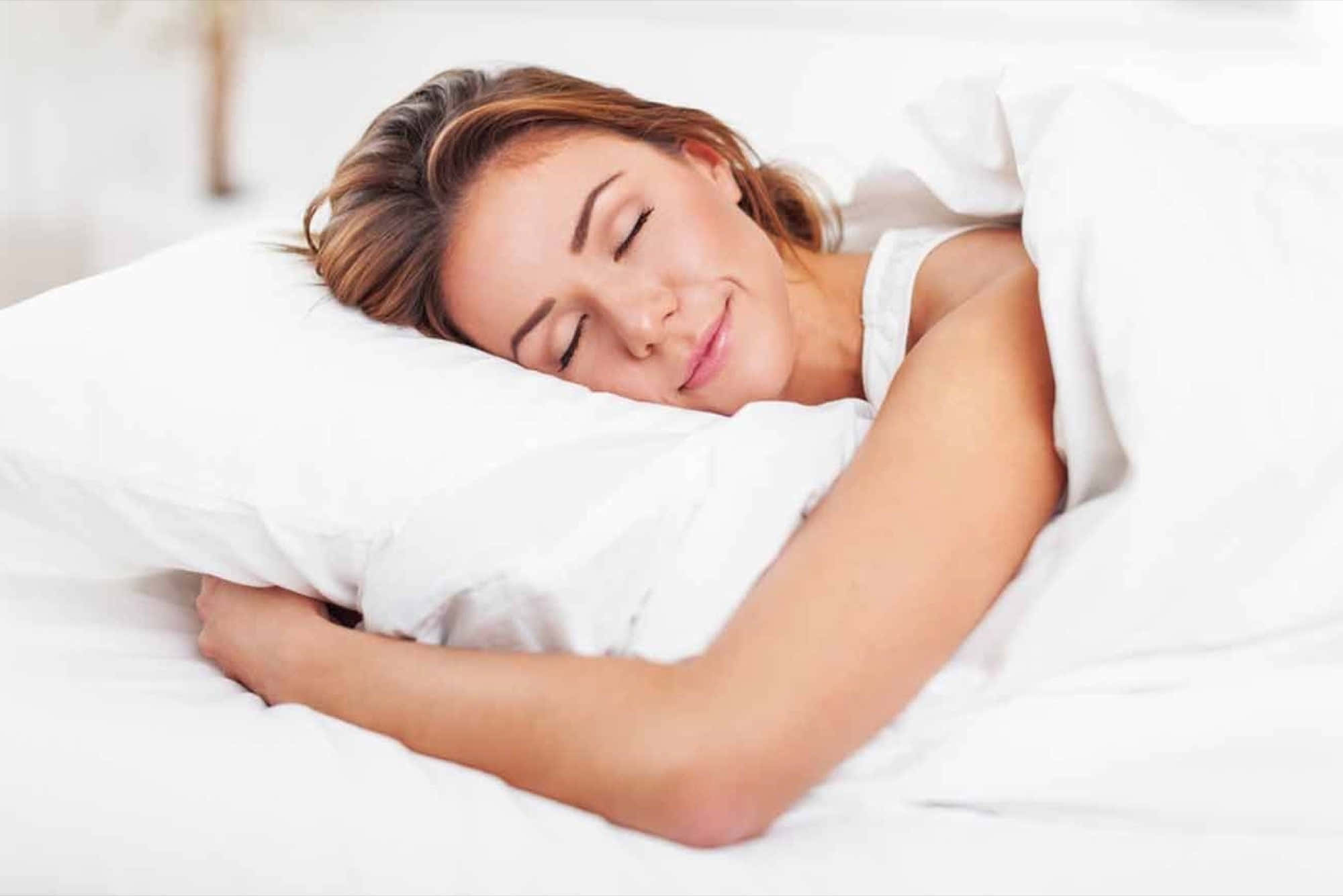 Sleep 桌布 壁紙 — 一位女士在明亮的臥室中，被柔軟的白色床單和枕頭包裹著，帶著溫柔的微笑安然入睡。