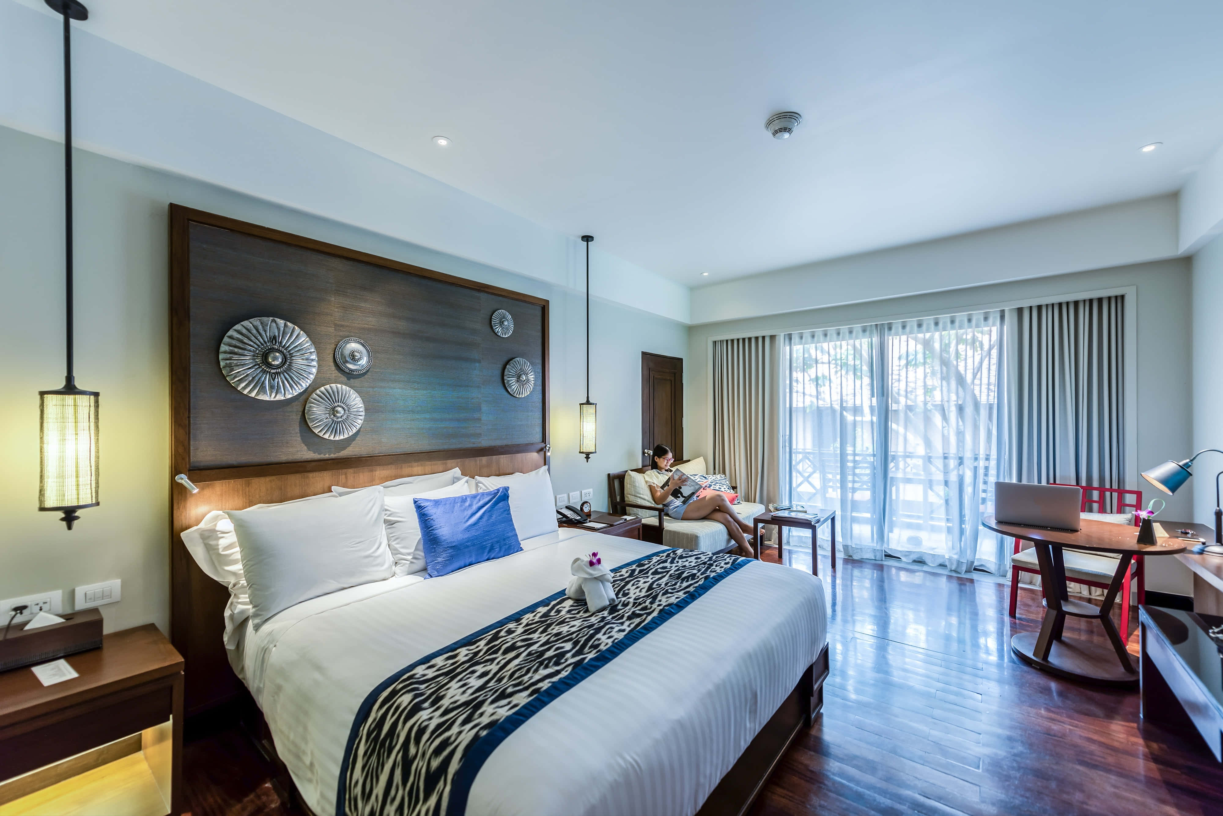 Hotel Room 桌布 壁紙 — 一位女士在現代酒店房間的沙發上放鬆，房間內有一張大床、木地板和落地窗，讓明亮的自然光透進來。