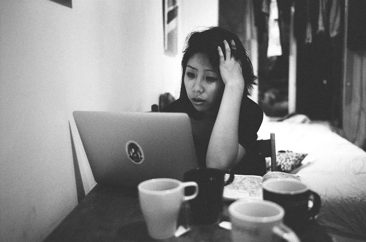 Working 桌布 壁紙 — 一位女士坐在一張雜亂的桌子旁，桌上放著多個杯子，在光線昏暗的房間裡專注地盯著她的手提電腦。