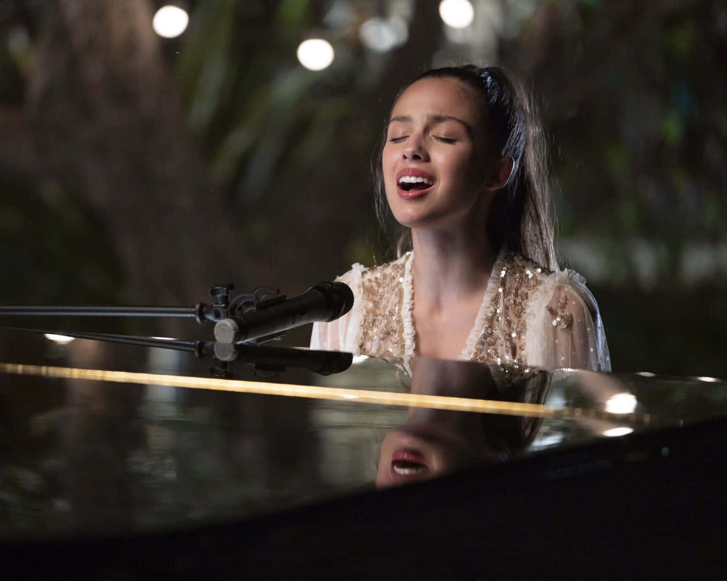 Olivia Rodrigo 桌布 壁紙 — 一位女士坐在三角鋼琴前，在柔和燈光下熱情地對著麥克風唱歌。