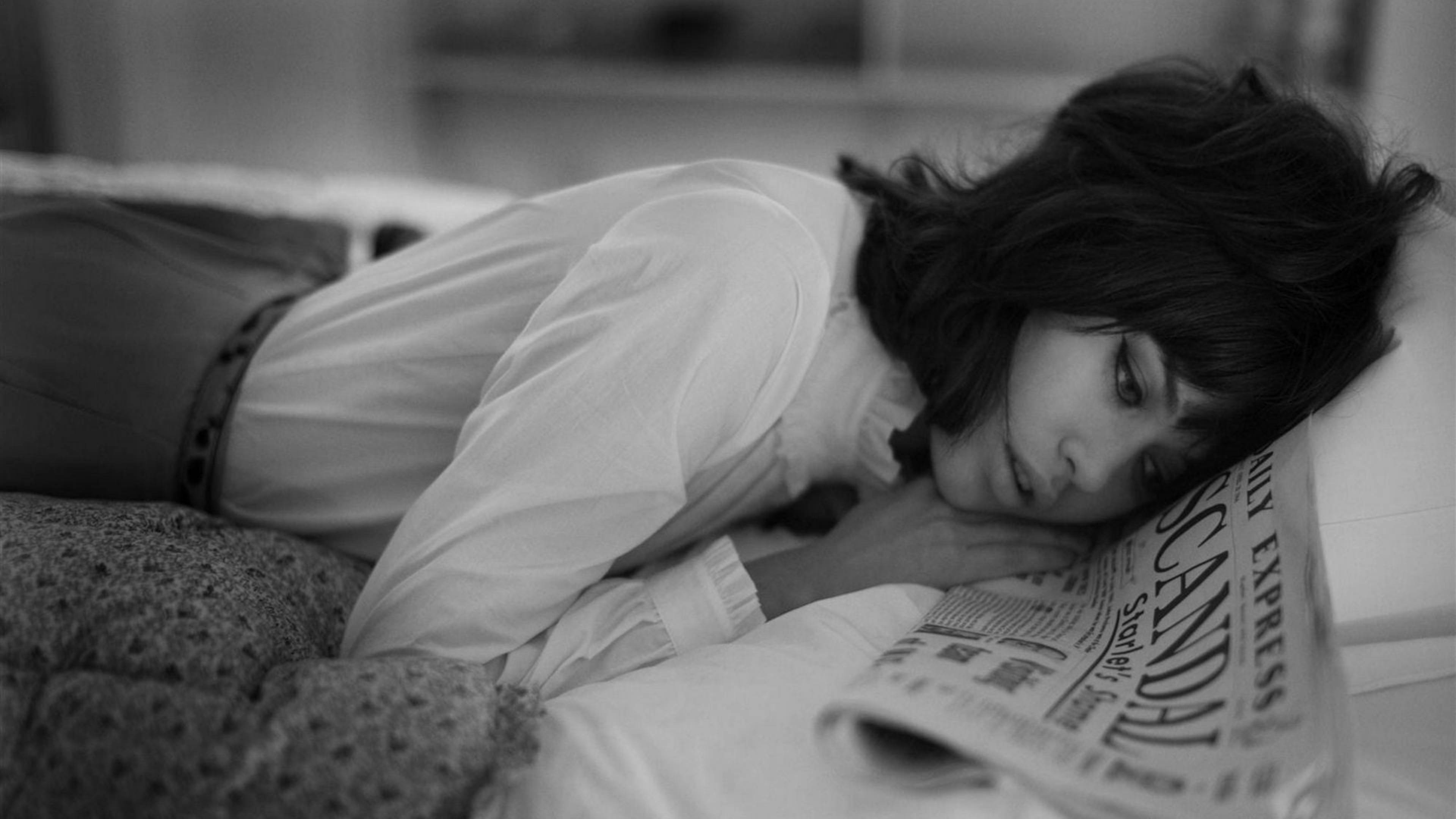 Newspaper 桌布 壁紙 — 一位女士將頭靠在枕頭上，在柔和光線的單色臥室裡閱讀報紙。