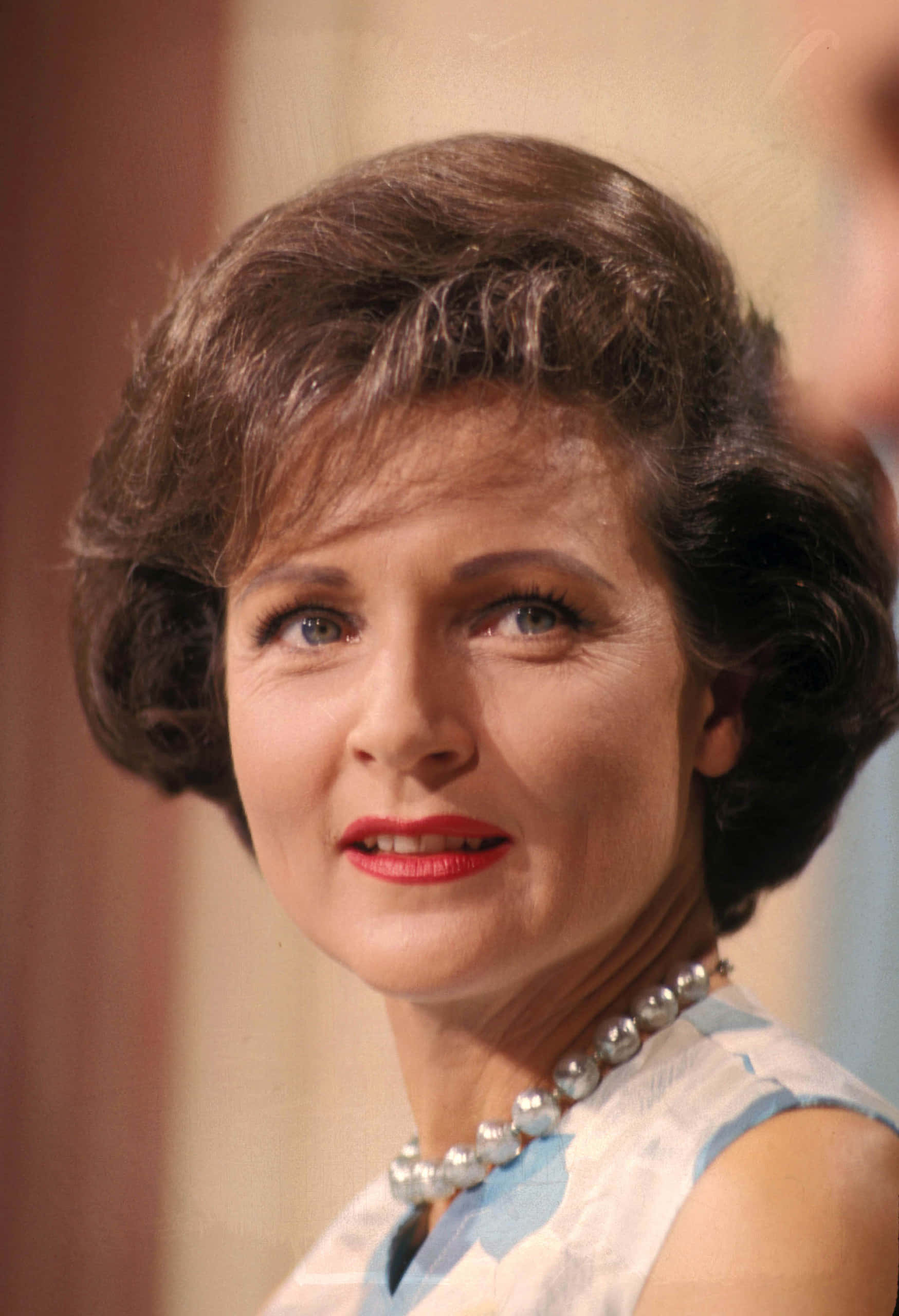 Betty White 桌布 壁紙 — 一位女士的復古肖像，她擁有造型棕色頭髮、紅唇和珍珠項鍊，向上凝視，帶著溫柔的微笑。