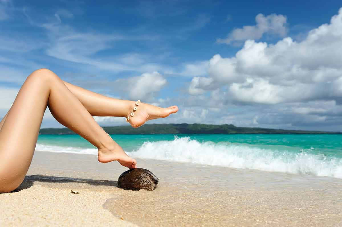 Jamaican Beach 桌布 壁紙 — 一位女士的赤裸雙腿擱在海岸線的椰子殼上，碧綠色的海浪在藍天白雲下拍打著岸邊。