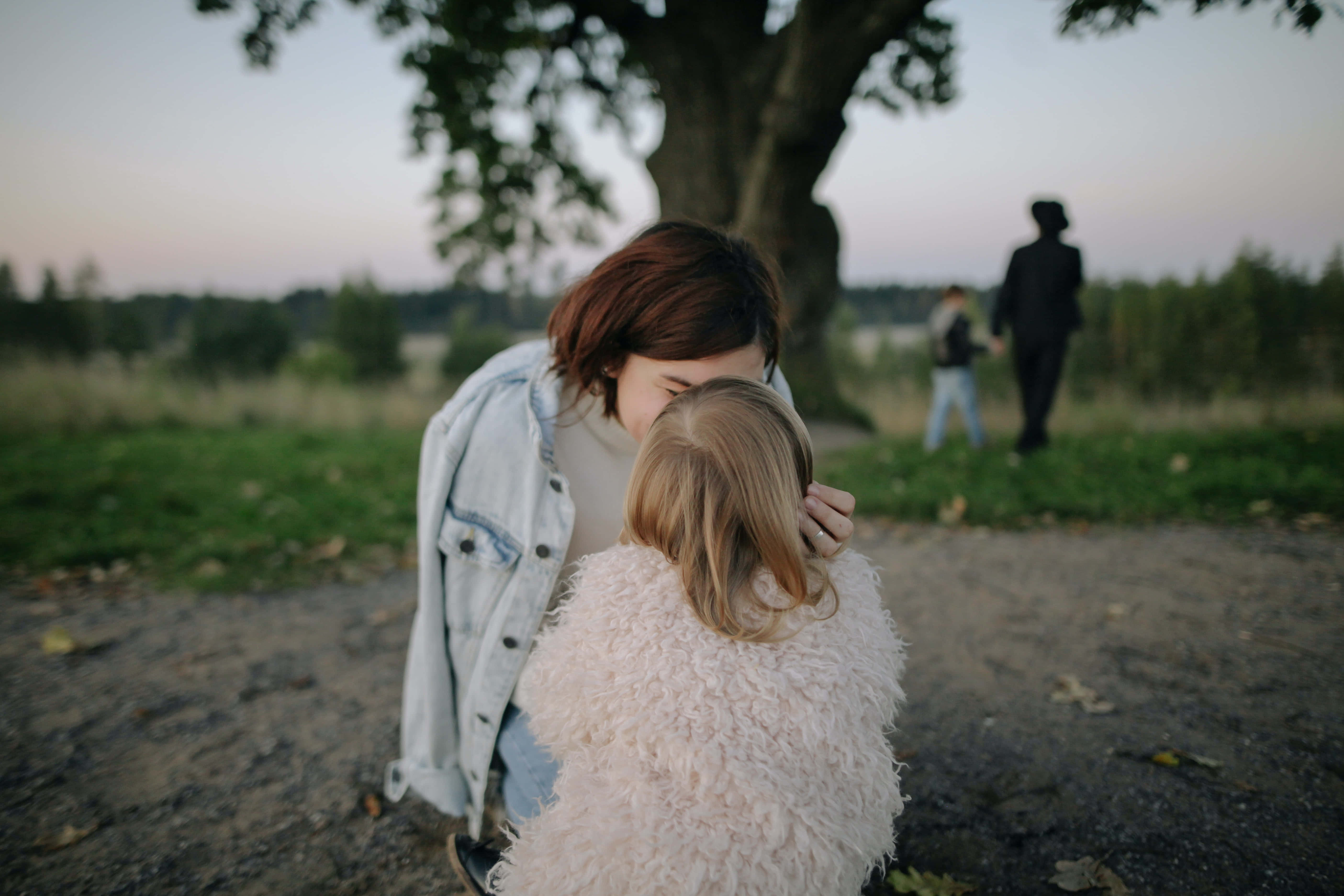 Mom 桌布 壁紙 — 一位女性在黃昏的草地上，在一棵大樹下溫柔地親吻一個孩子的頭。