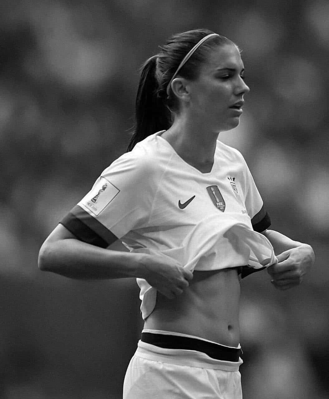 Alex Morgan 桌布 壁紙 — 一位女足球員在比賽中掀起球衣，露出結實腹部的黑白肖像。