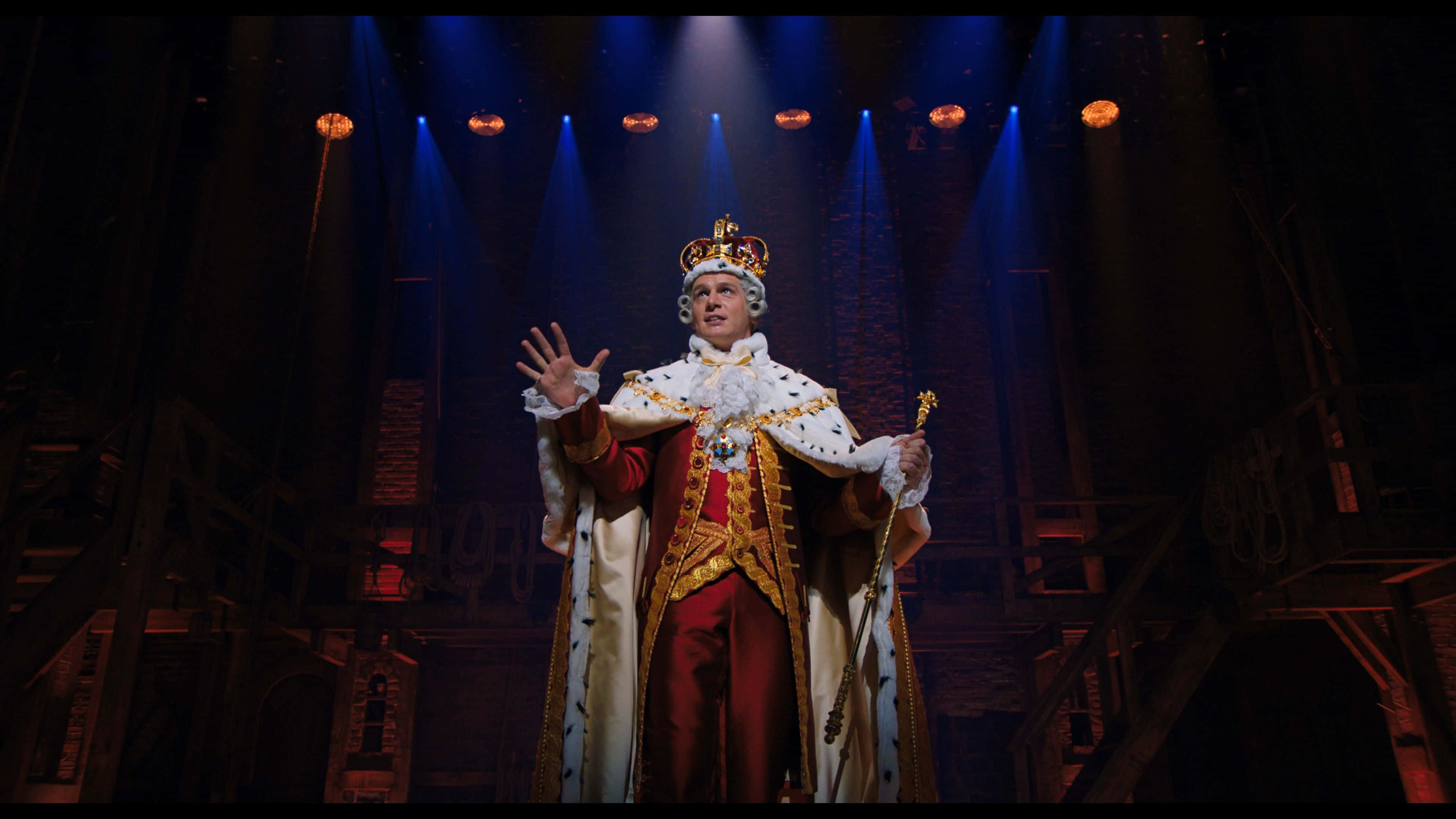 Hamilton 桌布 壁紙 — 一位威嚴的表演者戴著金色王冠和紅色長袍，在戲劇性的藍色和橙色聚光燈下掌控舞台。