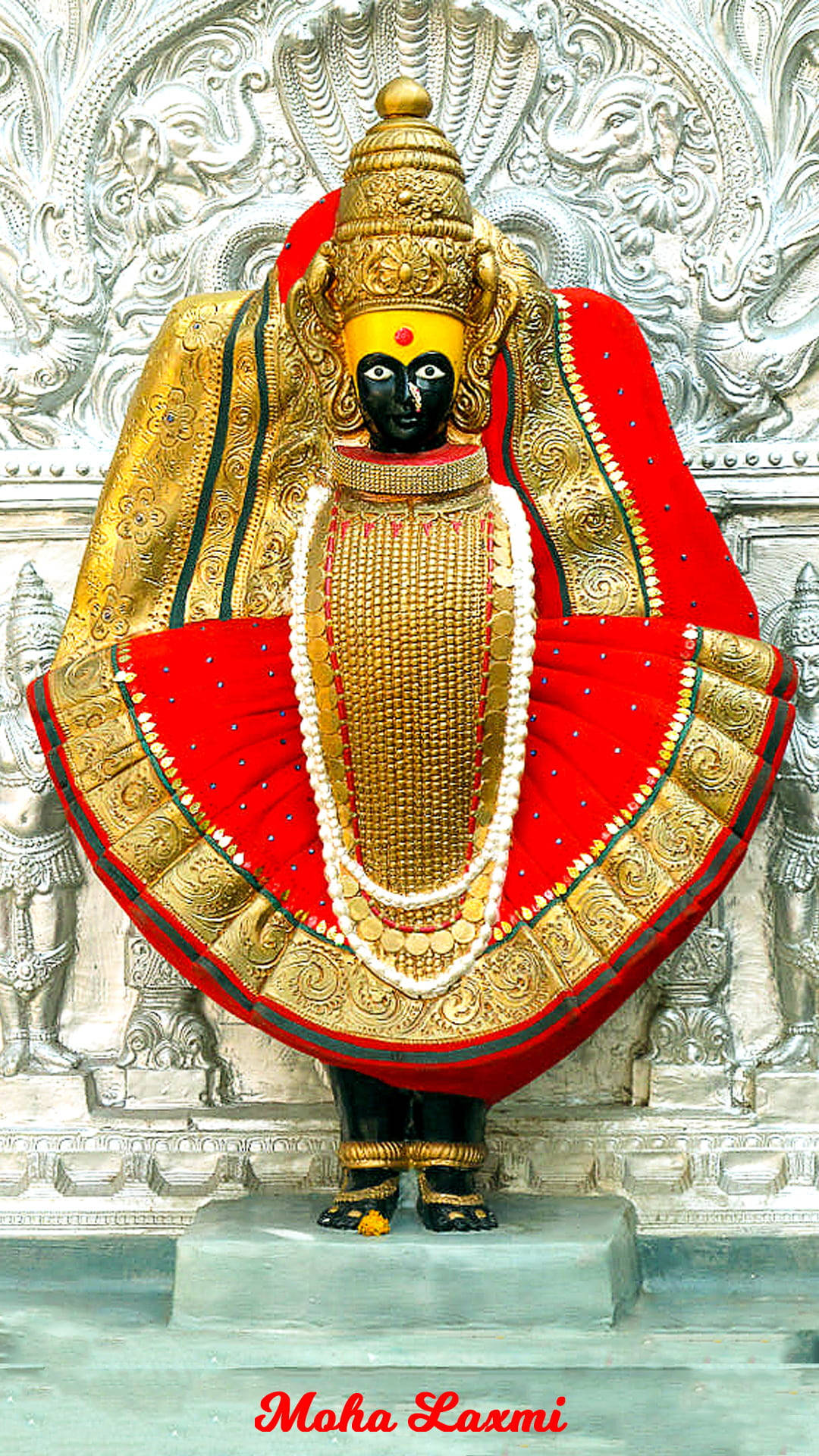 Lakshmi 桌布 壁紙 — 一位威嚴的黑面神祇，身著金色和紅色的禮服，站在雕刻精美的白色石廟背景前。