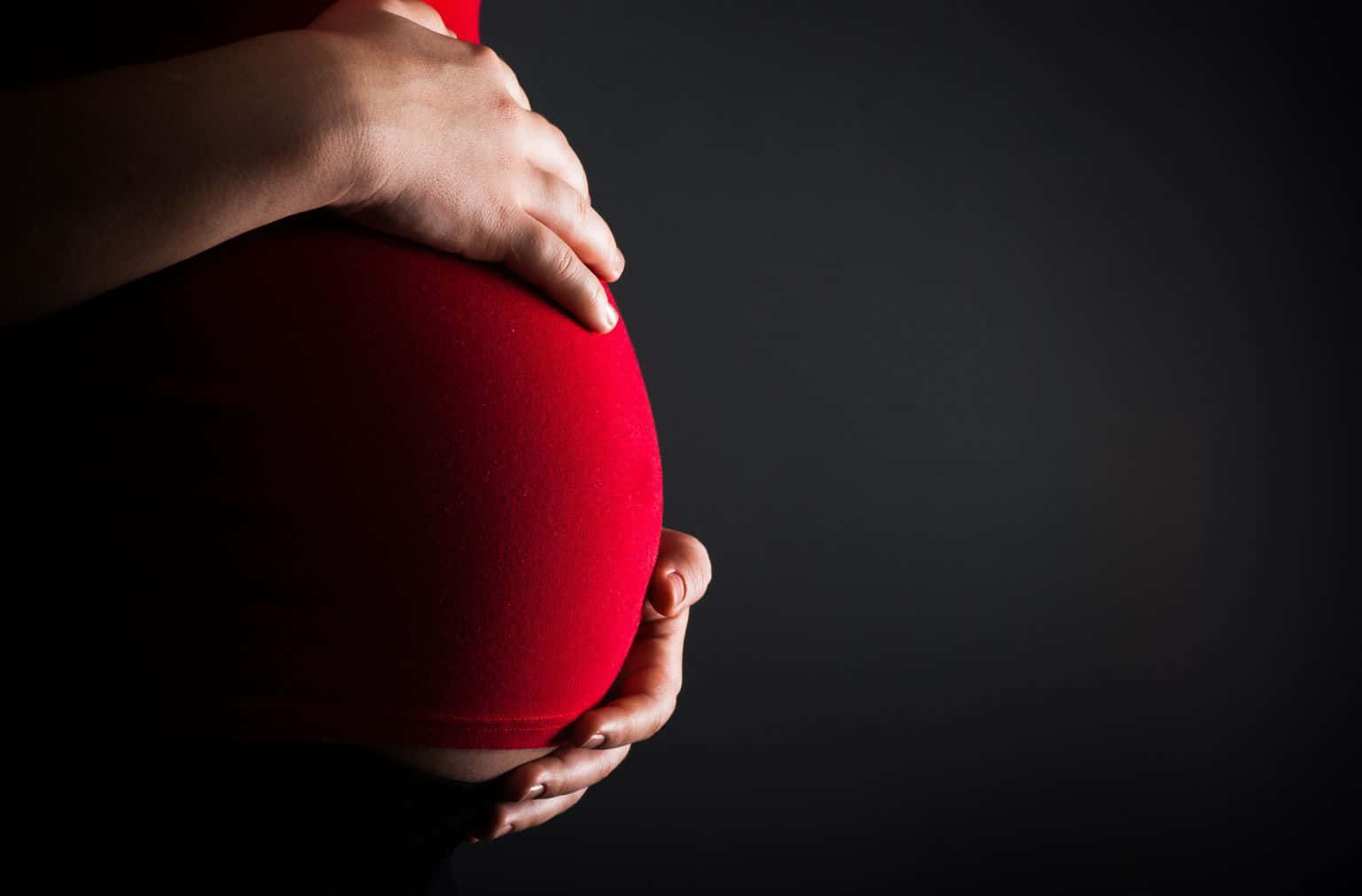 pregnancy 桌布 壁紙 — 一位孕婦的雙手在深色背景下捧著她的紅色孕婦上衣。
