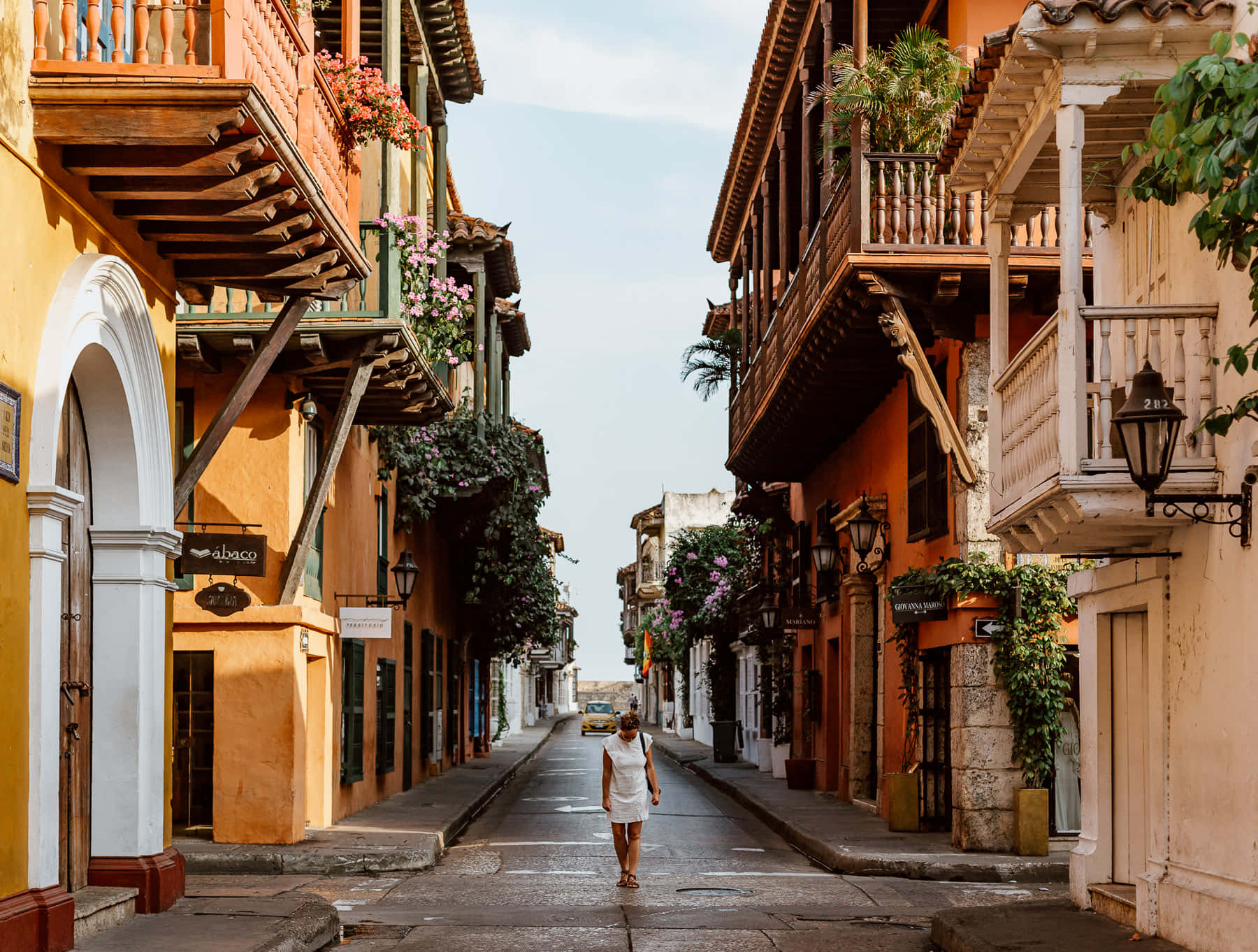 Colombia 桌布 壁紙 — 一位孤獨的女人走在陽光普照的殖民風格街道上，兩旁是色彩繽紛的建築物和懸掛的花朵。