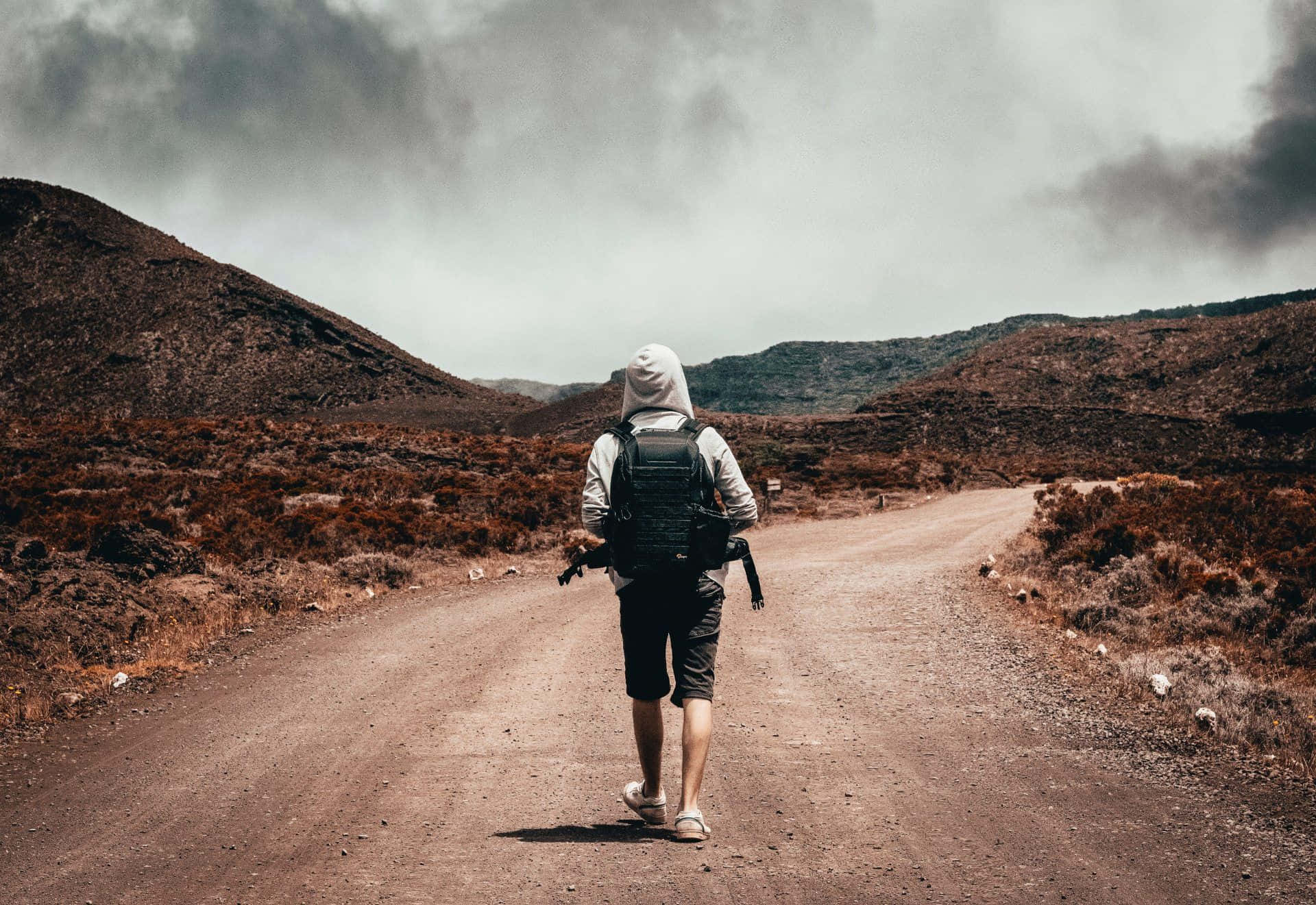 Hiking 桌布 壁紙 — 一位孤獨的揹著背包的遠足者，在陰天之下，沿著一條蜿蜒的泥路穿過一片貧瘠、岩石遍地的景觀。