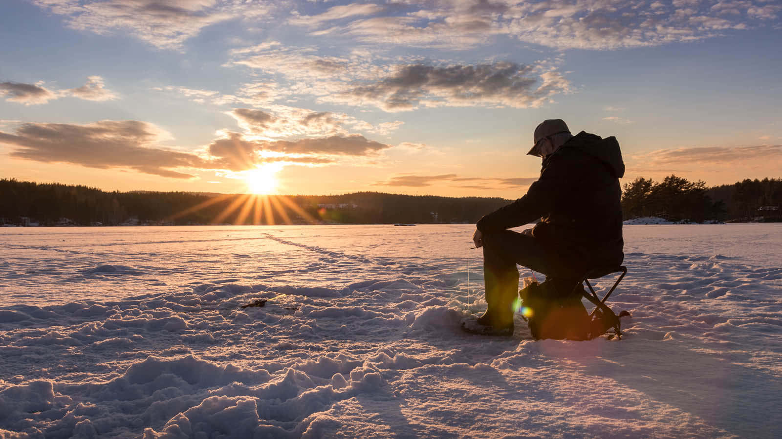 Ice Fishing 桌布 壁紙 — 一位孤獨的漁夫在日落時分坐在結冰的湖面上的凳子上，在金色天空和散落的雲朵映襯下形成剪影。