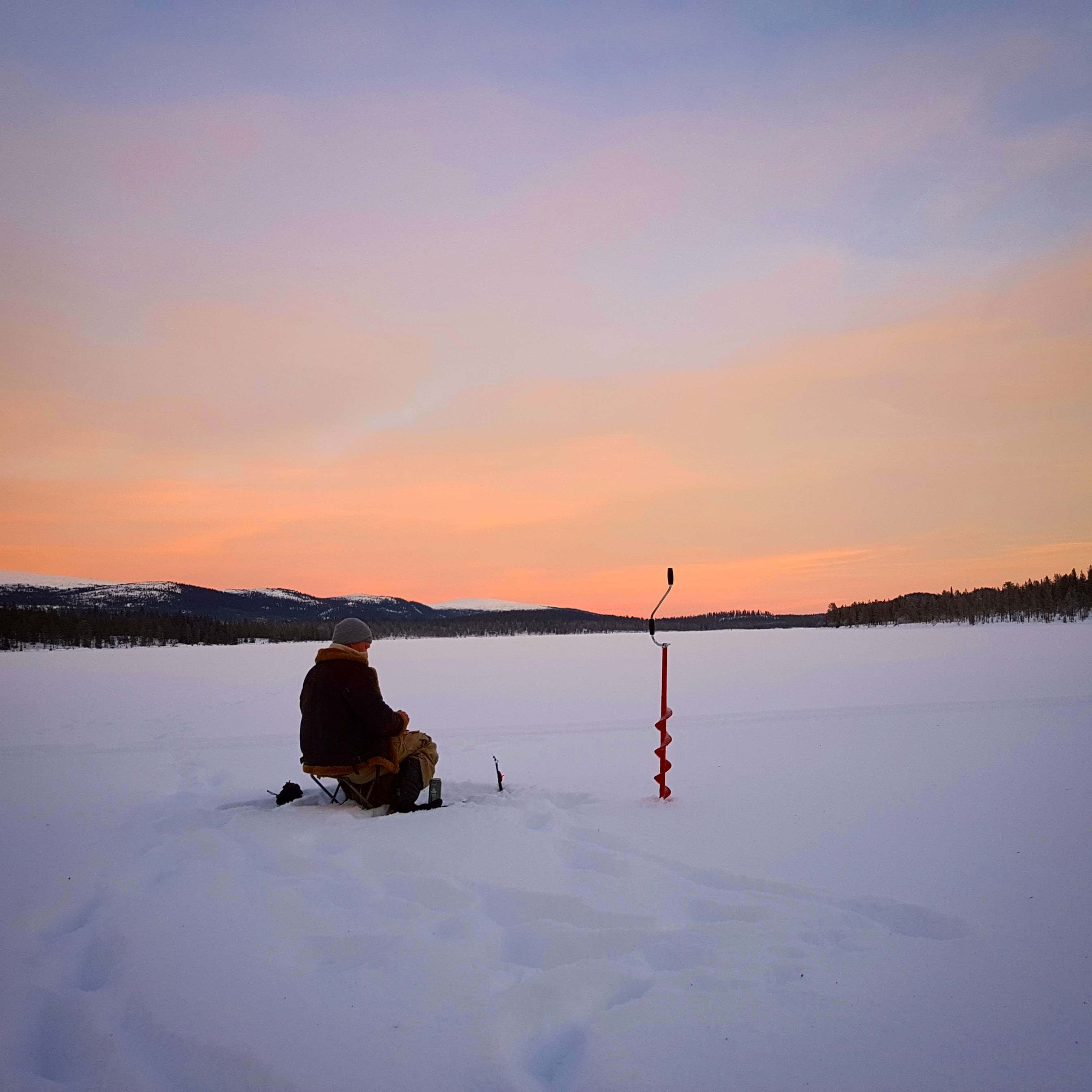 Ice Fishing 桌布 壁紙 — 一位孤獨的漁夫坐在白雪覆蓋的冰凍湖面上，夕陽西下，身旁立著冰鑽，遠處白雪皚皚的山丘在柔和天空下。