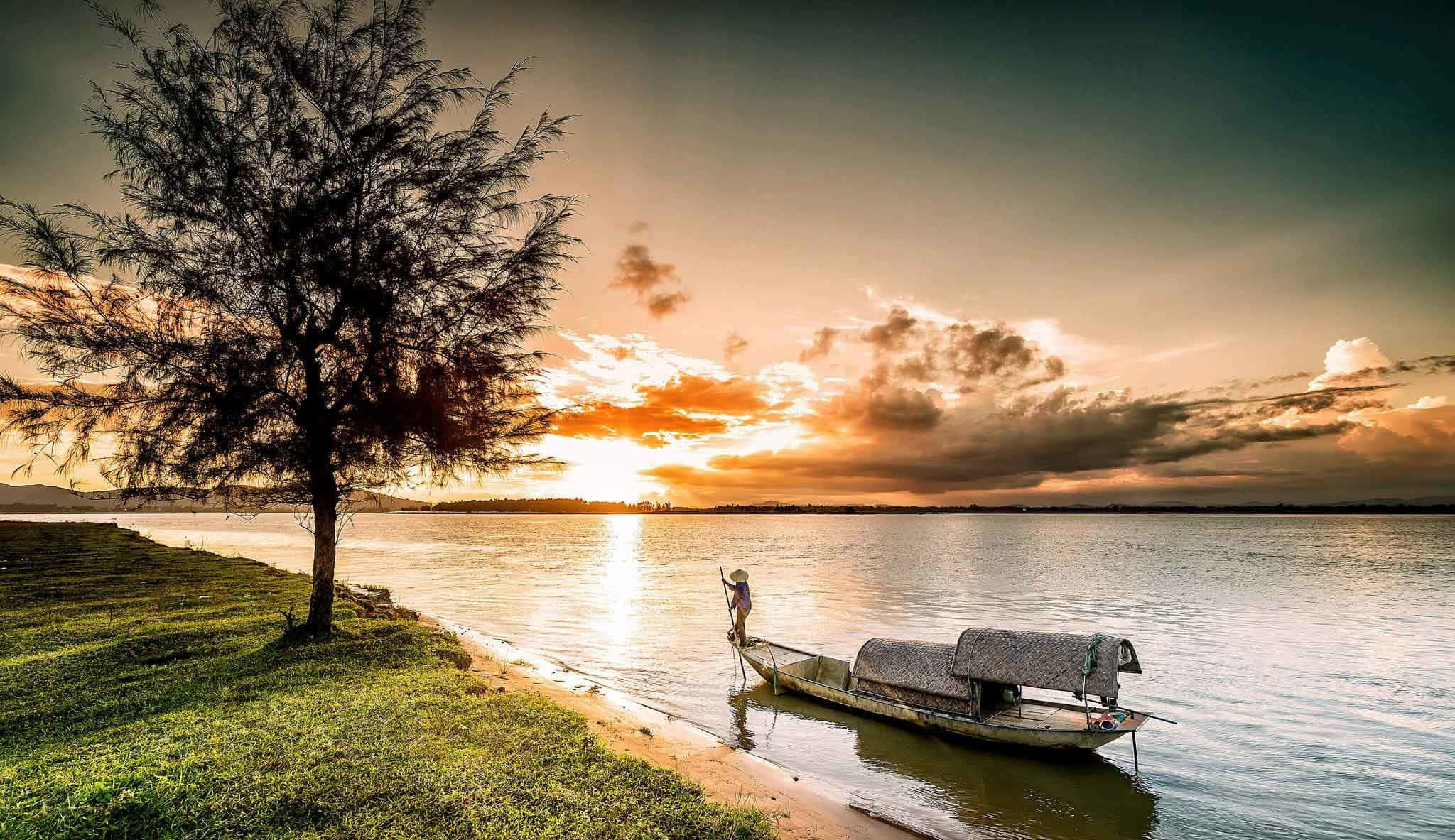 Vietnam 桌布 壁紙 — 一位孤獨的漁夫站在木船上，夕陽西下，在火紅的天空和寧靜的水面上形成剪影，旁邊是長滿草的岸邊，有一棵孤樹。