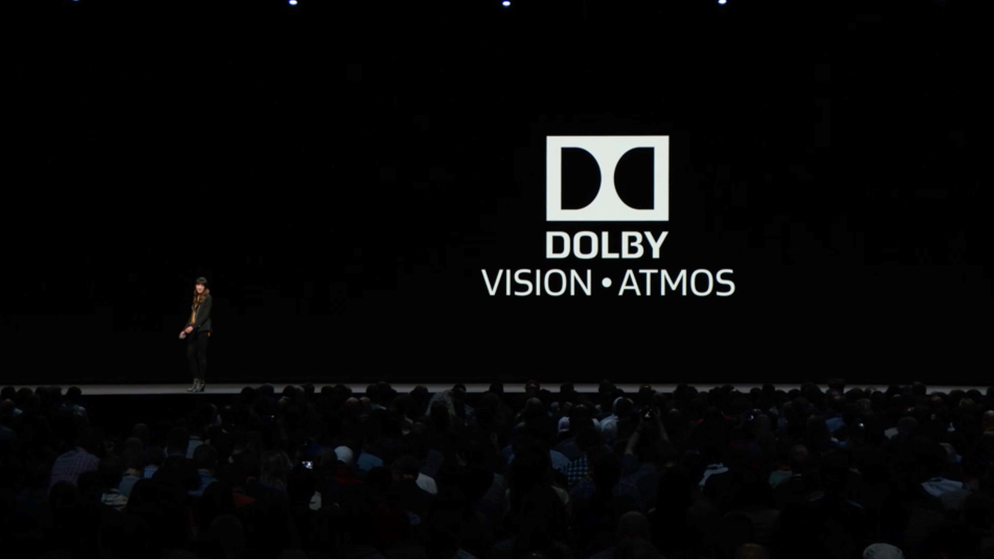 4K Dolby Vision 桌布 壁紙 — 一位孤獨的演講者站在黑暗的舞台上，面對著一個巨大的屏幕，屏幕上顯示著Dolby Vision Atmos標誌，面向一片剪影般的觀眾。