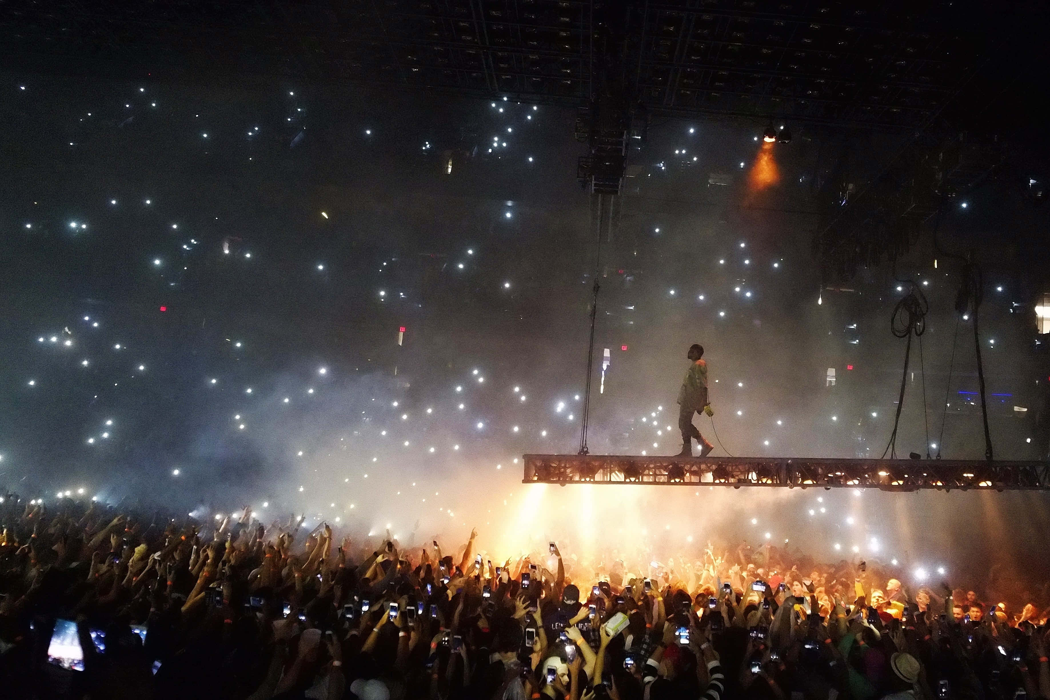 Kanye 桌布 壁紙 — 一位孤獨的表演者在燈光照耀的舞台上行走，下方是如夜空繁星般舉起發光手機的粉絲海洋。