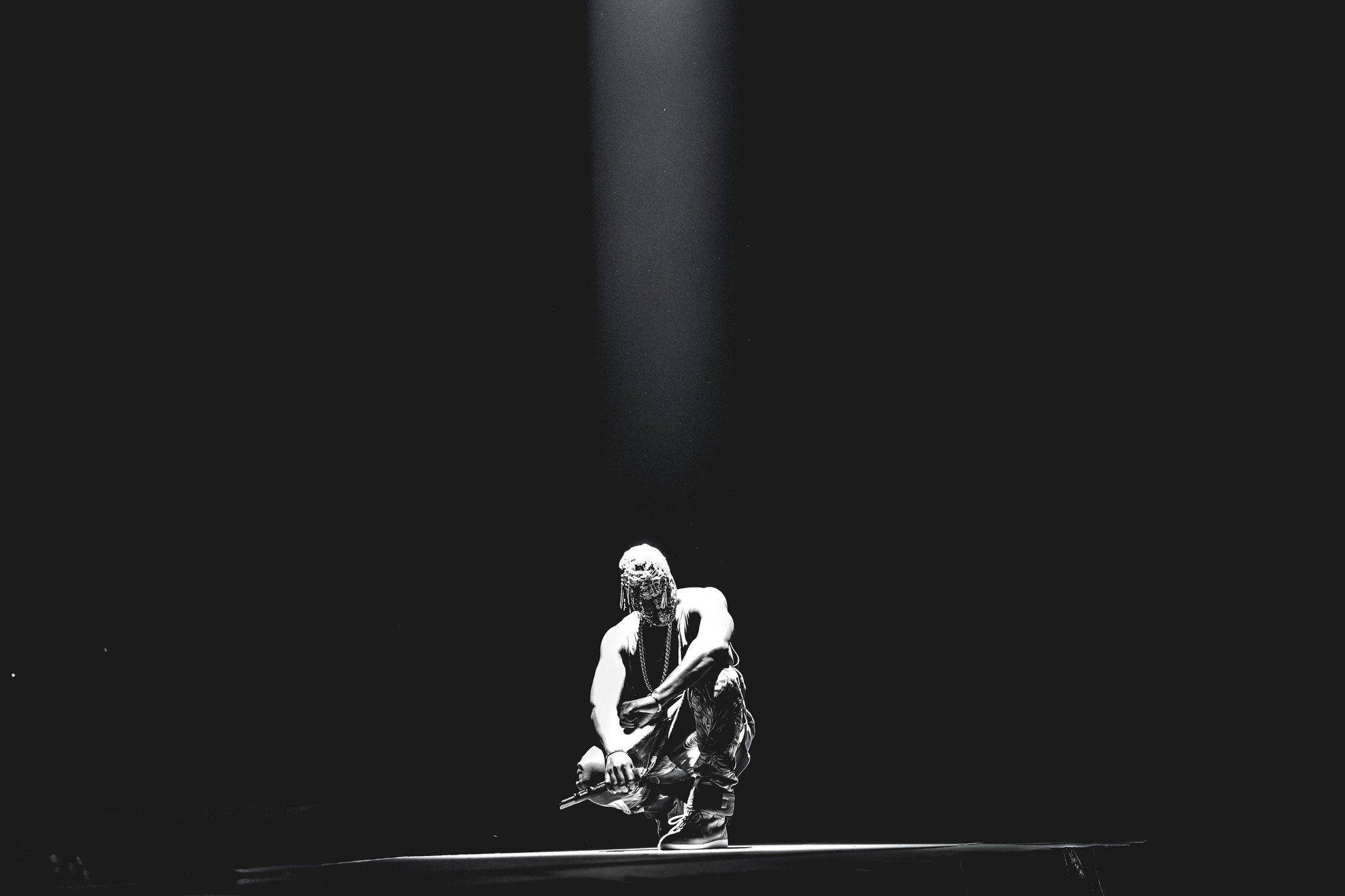 Kanye West Kneeling Under Light 壁紙 — 一位孤獨的表演者在黑暗的舞台上蹲在單一聚光燈下，營造出戲劇性和強烈的氛圍。