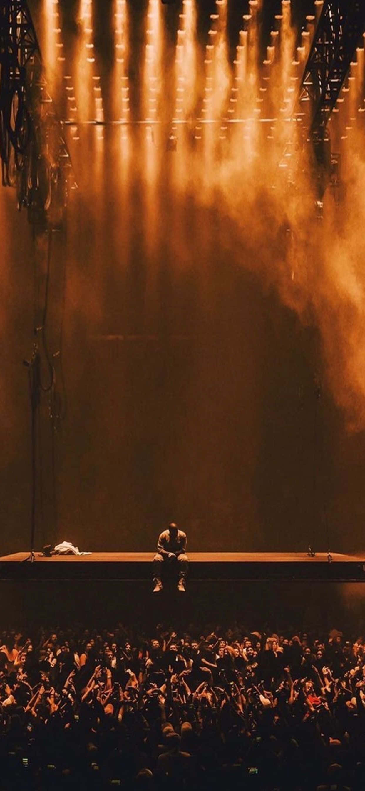 Kanye 桌布 壁紙 — 一位孤獨的表演者坐在舞台中央，沐浴在金色聚光燈下，下方人山人海的粉絲伸手向上。