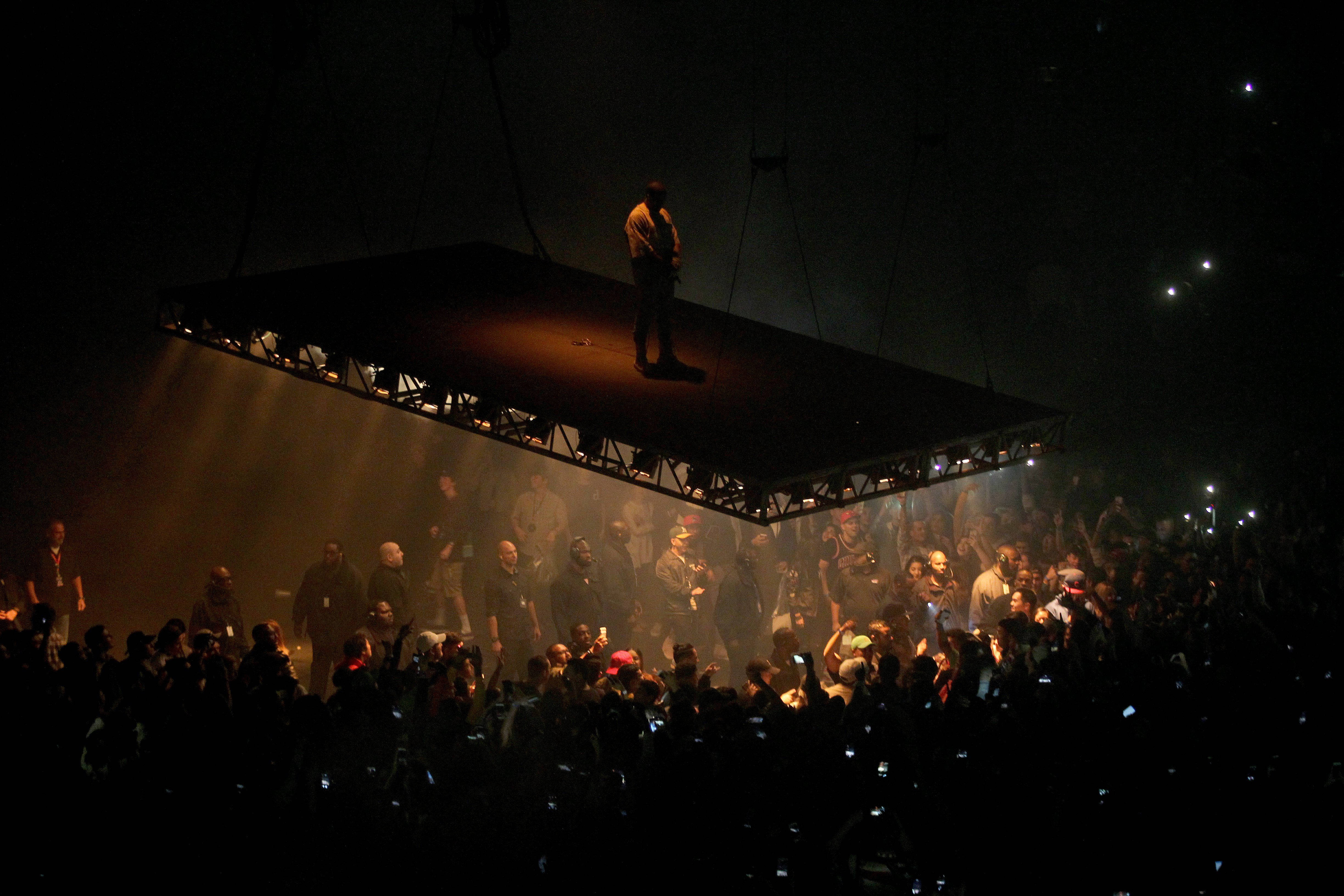 Kanye West On Elevated Stage 壁紙 — 一位孤獨的表演者獨自站在懸浮舞台上，下方是煙霧彌漫的音樂廳中舉著發光手機的粉絲海洋。